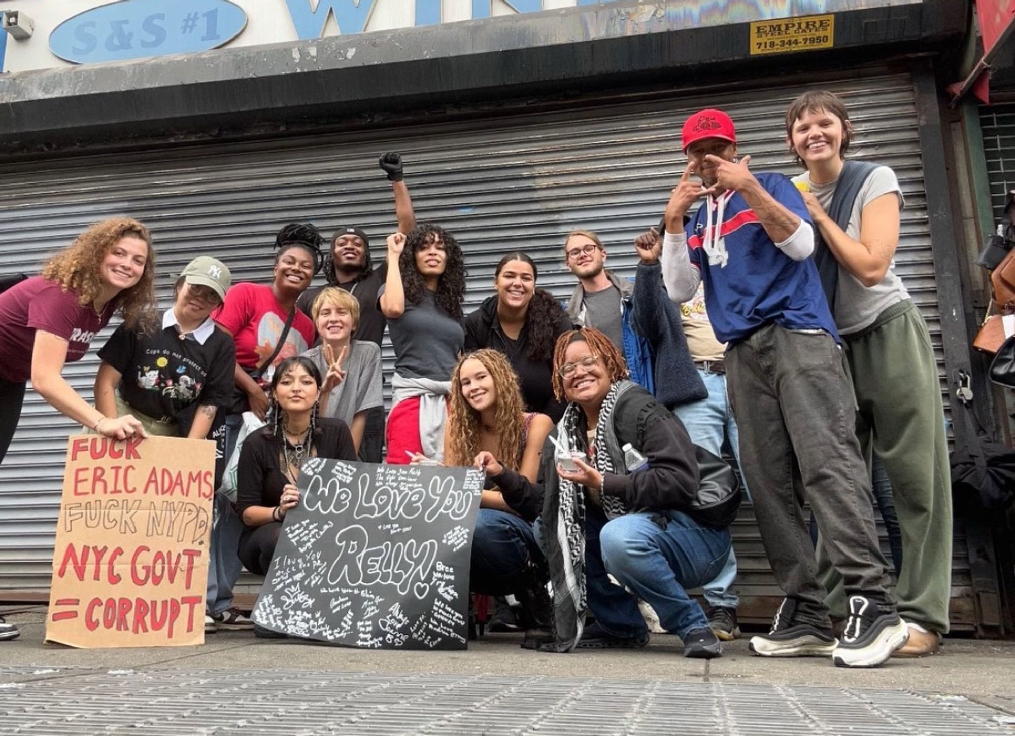 We The People members after distro posing for a group photo with signs that say “we love you Relly” with handwritten messages and another that says “fuck Eric Adams, fuck NYPD, NYC Govt=Corrupt”