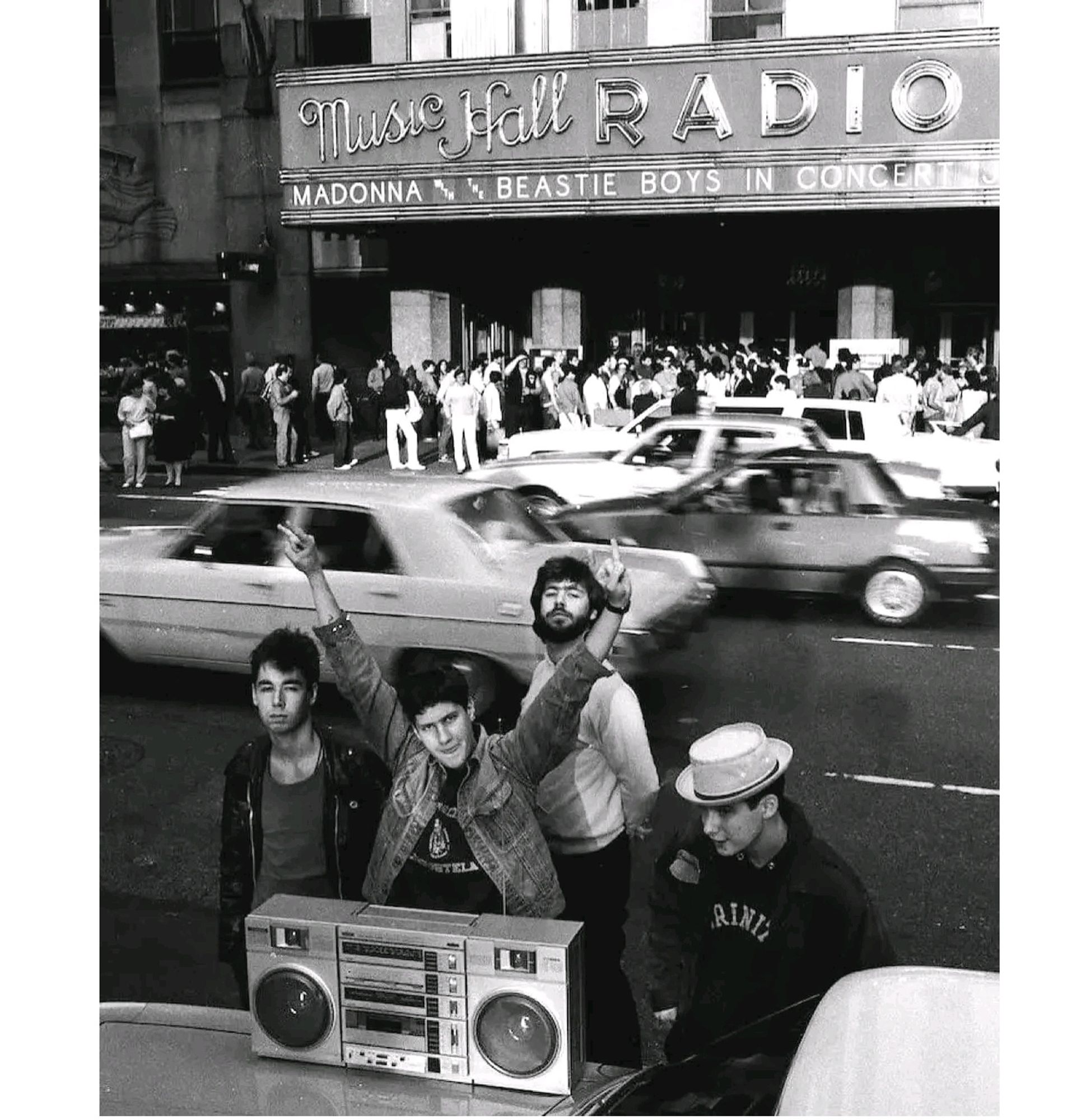 Foto der Band Beastie Boys. 4 Musiker stehen mit einem Ghettobluster im Vordergrund. Hinter ihnen fahren unscharf Autos, dahinter sieht man eine Menschen Menge vor einem Gebäude mit der Aufschrift 'music hall radio, Madonna beastie Boys in concert'