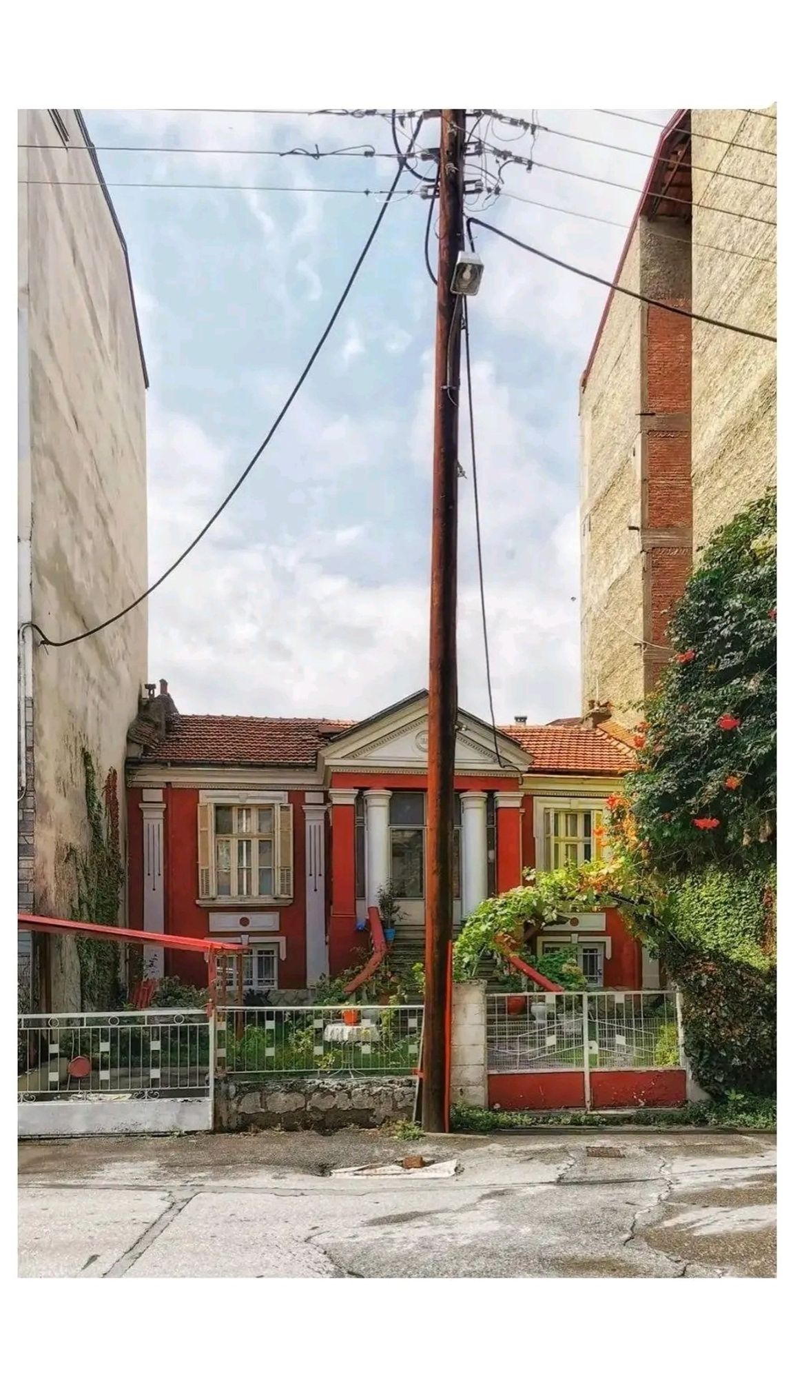 Ein kleines altes Haus mit Veranda, eingeklemmt zwischen zwei großen. Darüber blauer Himmel