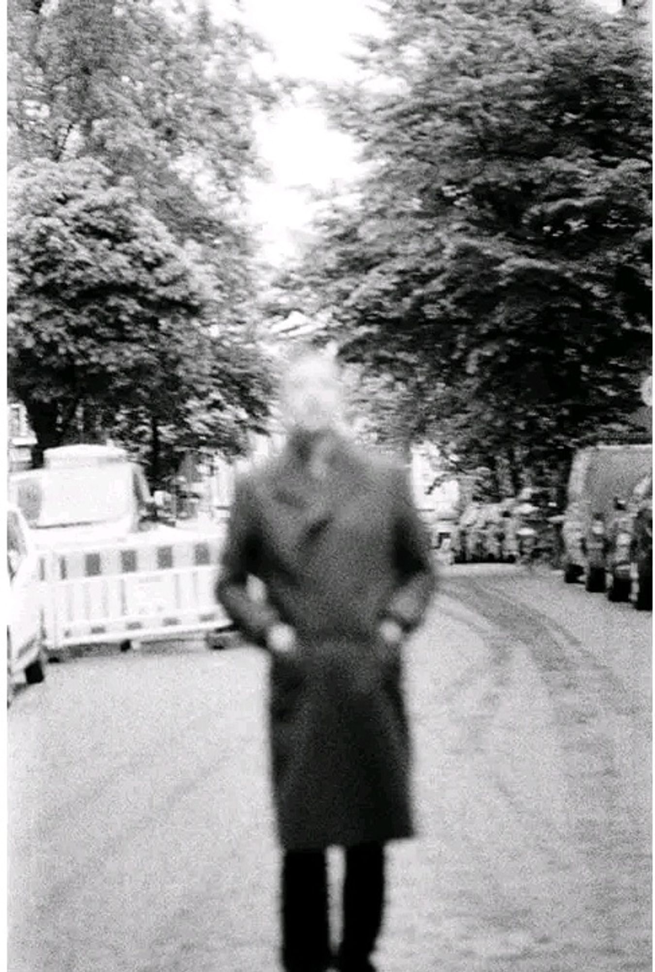 Körniges #sw #Foto ein Mann in einem Mantel im Vordergrund in der Mitte einer Straße. Das Gesicht ist unscharf. Im Hintergrund links ein bauzaun, darüber dichte Baumkronen. Rechts sieht man parkende Autos