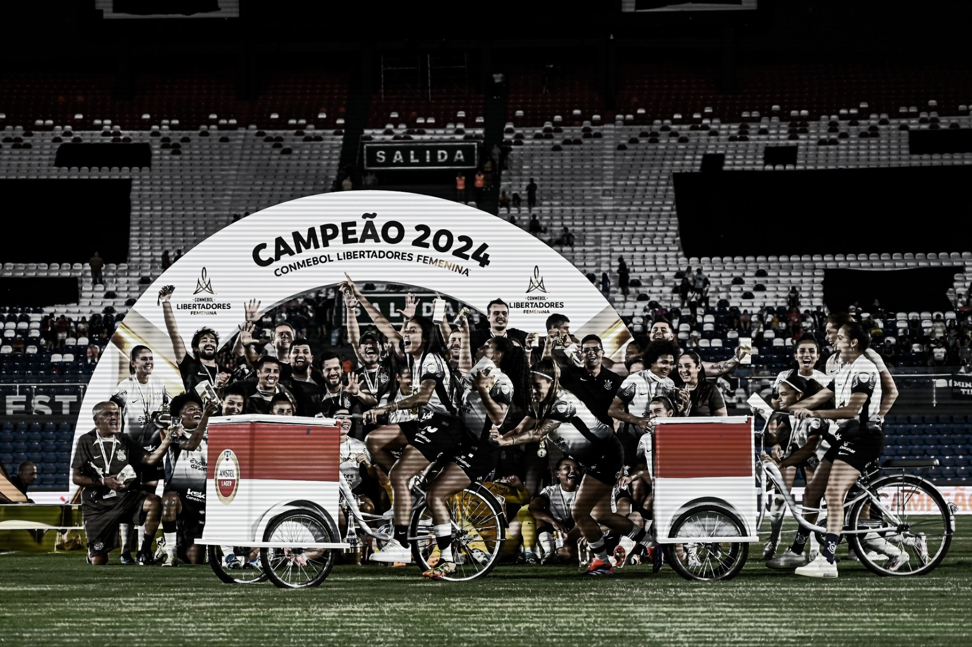 Foto de todo o elenco e comissão técnica do Corinthians comemorando no palco de Campeão 2024 da Libertadores Feminina. Algumas jogadoras felizes andam com a bicicleta do patrocinador Amstel (e os respectivos isopores com cerveja) no primeiro plano.
Créditos: Staff Images Woman / CONMEBOL