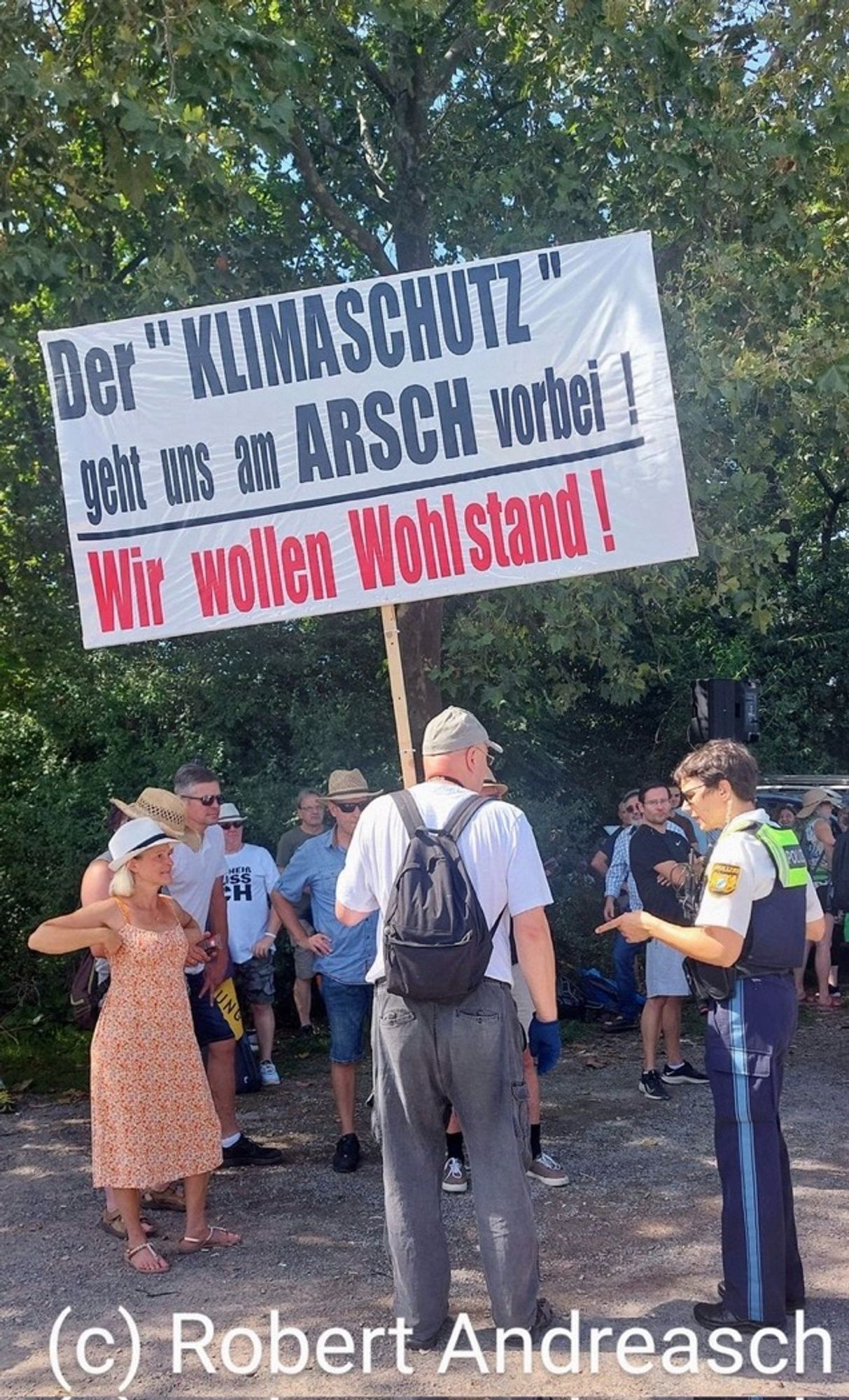 Foto eines alten Mannes in Diskussion mit einer Polizistin. Er hält ein Plakat mit der Aufschrift: Der Klimaschutz geht uns am Arsch vorbei! Wir wollen Wohlstand!
Im Hintergrund stehen Menschen vor grünen Bäumen