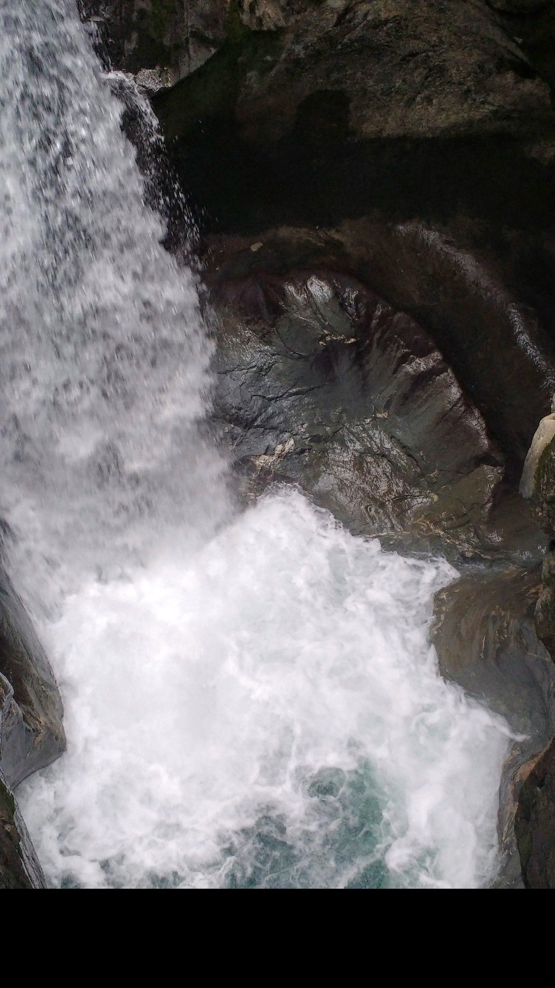 Ein tosender Wasserfall schießt von der linken Seite nach unten in ein vom Wasser ausgespültes Becken, weiße Gischt, Wasserstrudel und dunkles Felsgestein