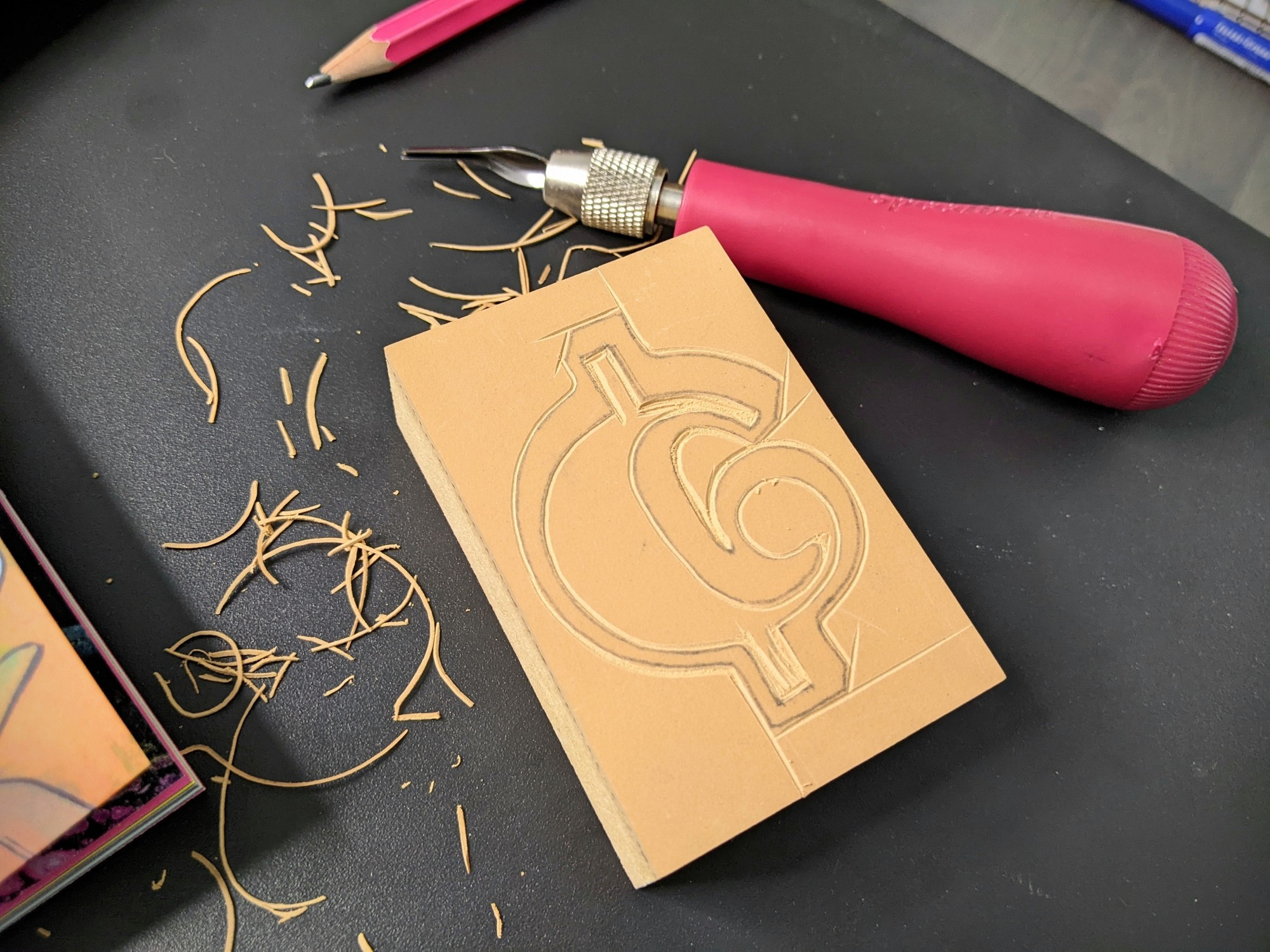 A partially carved linoleum block for printing sits next to a carving tool and pile of linoleum shavings. The design being carved is a decorative ¢ sign.