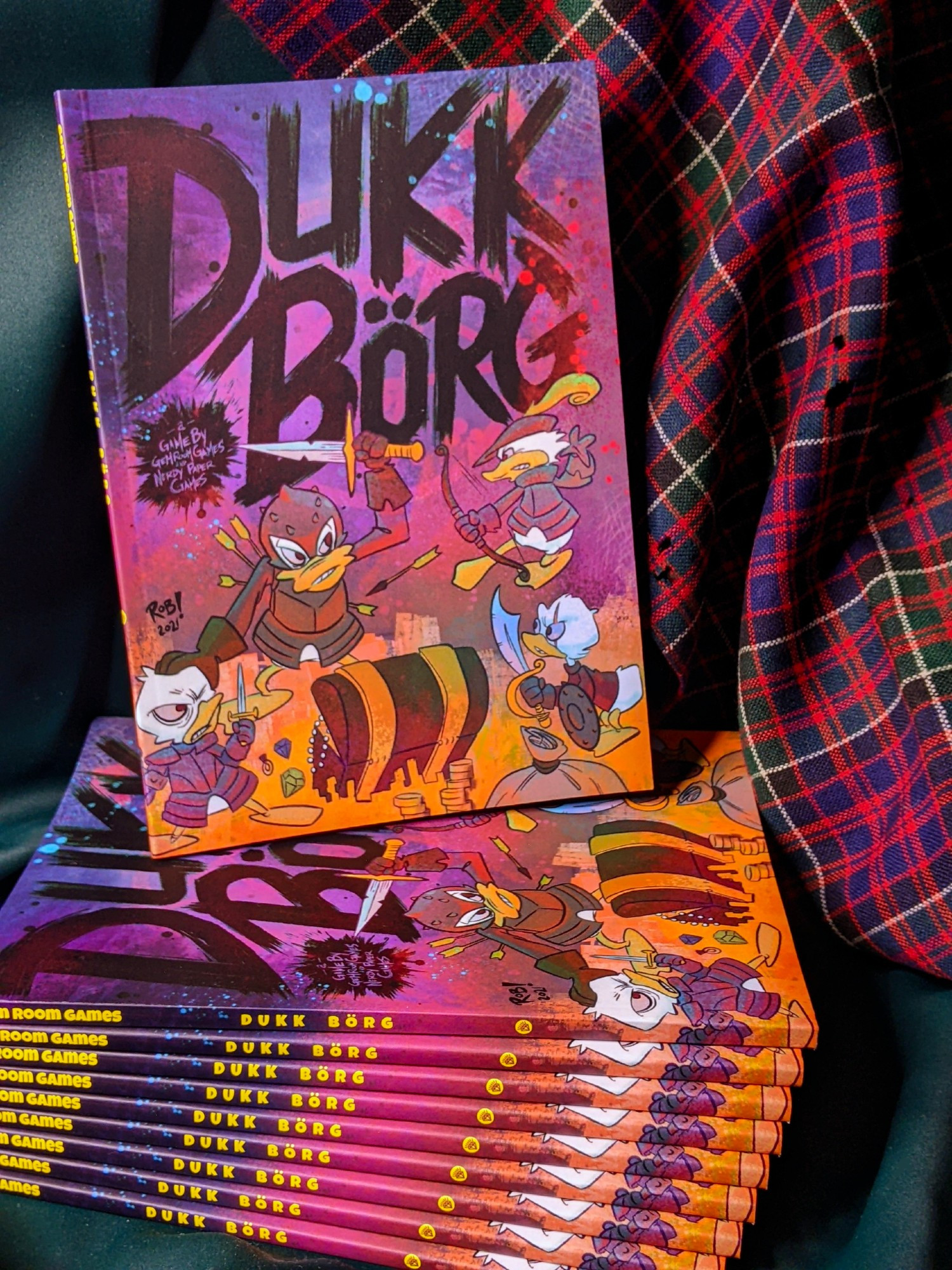 A stack of DUKK BÖRG proofs fanned on their side to show their spines with one copy upright on top to show the cartoon ducks in battle on the cover. The pile rests against dark green cloth and a moth-eaten red tartan.