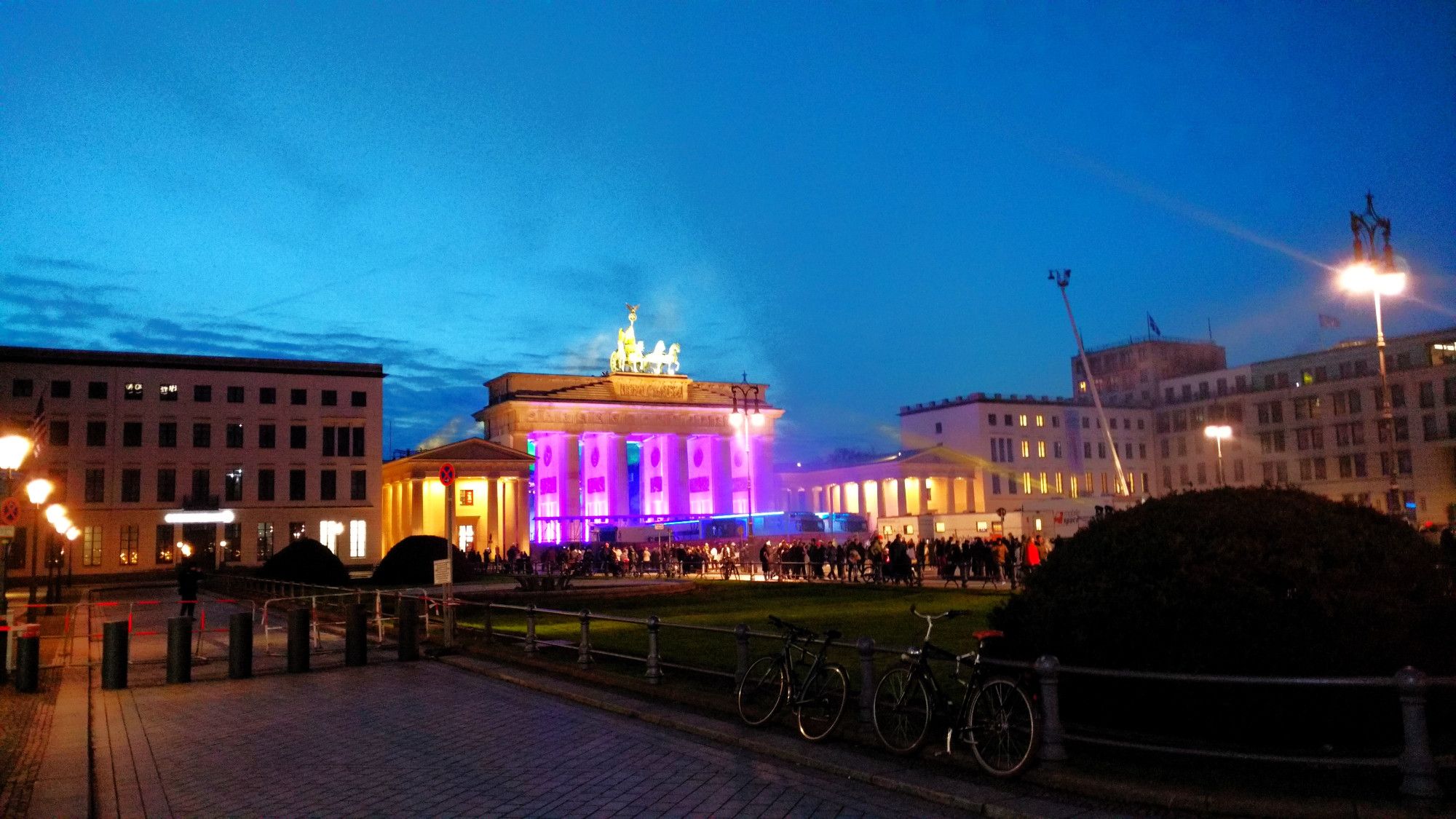 Auf dem Foto der Pariser Platz und das Brandenburger Tor. Das Tor ist lila und gelb beleuchtet, davor einige Menschen. Der Himmel ist dunkelblau, die Laternen sind an, es ist dämmrig.