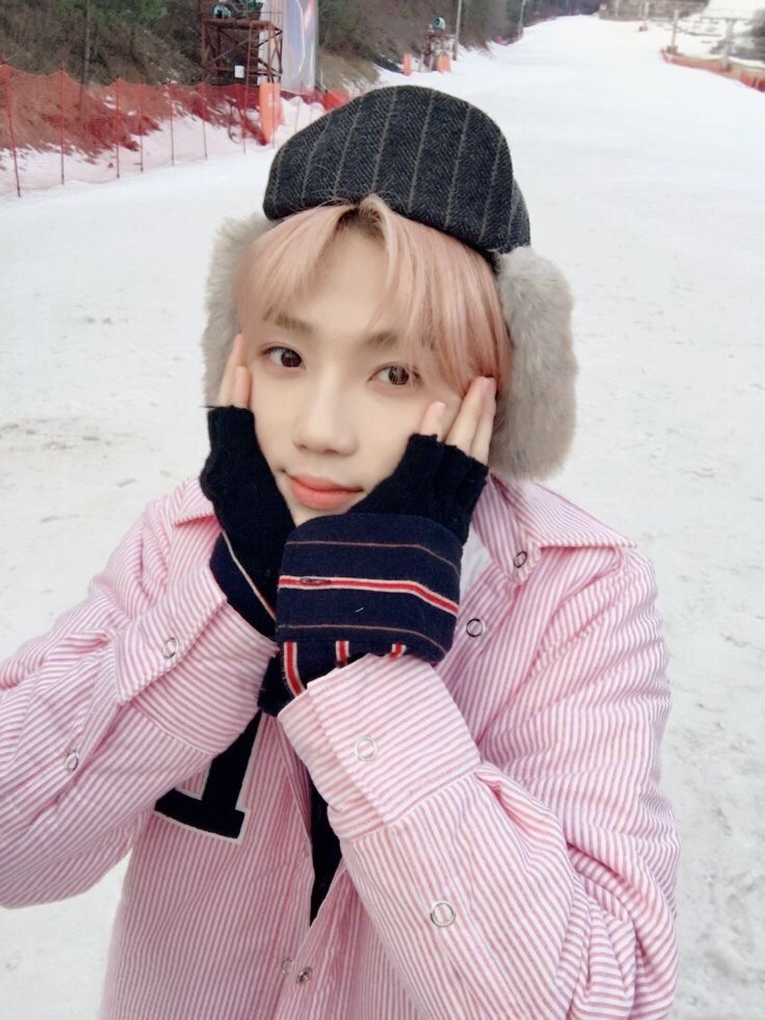 chanhee aka new with light pink hair, holding his face in both hands and looking at the camera. he's wearing a hat and the background is snowy