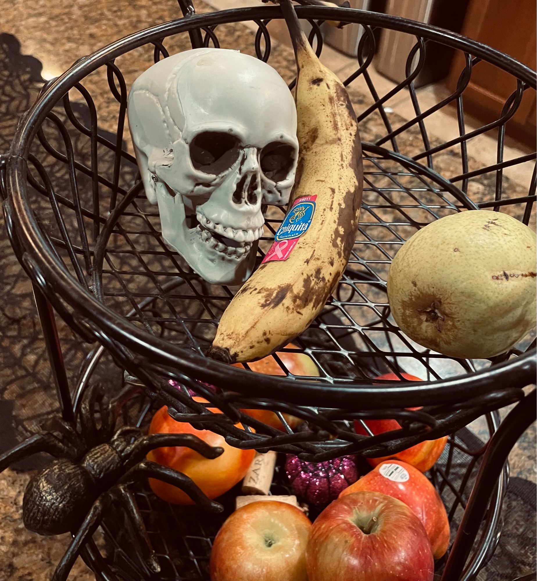 A black, woven metal fruit basket containing several pieces of fruit, a metal spider, and a plastic skull that is positioned to look like it's talking on a banana like a phone.