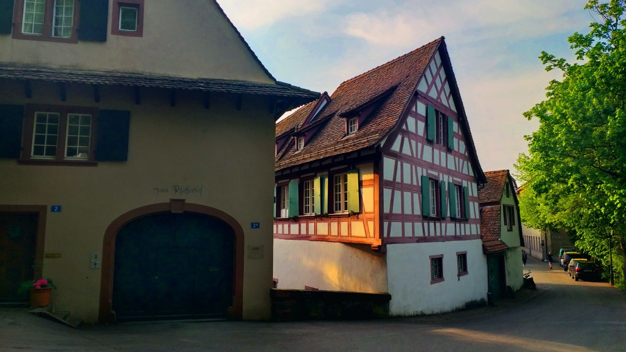 der abendhimmel ist leicht bewölkt mit sonnenschein. rechts ragt eine blühende baumkrone ins bild. schattige hausfassaden ziehen sich durch die szene. heraus ragt der fachwerkbau des schindelhofs, dessen längsseite in der abendsonne leuchtet