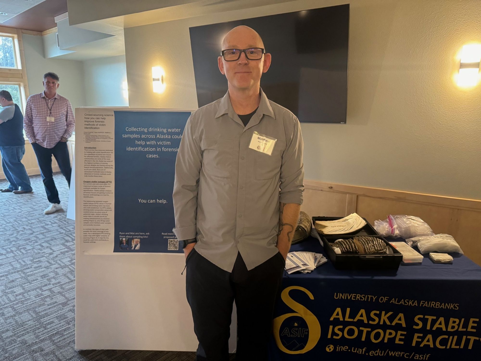 Professor Mat Wooler, a white man with glasses, standing in front of an academic poster and a table labeled 'Alaska Stable Isotope Facility'