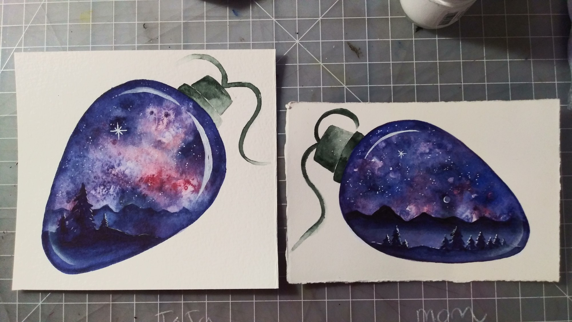 Two watercolor holiday cards on a gray cutting mat with white grid markings. Each card has a white background, with a painting of a clear C-7 Christmas light bulb. The bulbs are elongated and wider at the top, where a green cap shows a short bit of the cord that would make up the light string. Inside, each bulb is a small landscape scene featuring a fantastic nebula sky. Soft reds and violets mingle among deep indigo spaces and many stars. At the horizon, each scene has a distant mountain range in silhouette against the sky. In the foreground, we see a silhouette of a few pine trees, backlit by the brilliant stars. Painted highlights remind us we are looking into a glass bulb. Painted with watercolor and gouache on 140lb watercolor paper. The left card is approx. 5" x 5" and the right is 4" x 6".