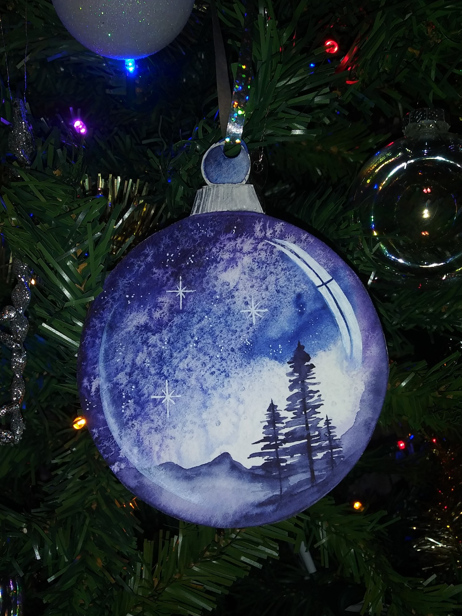 A watercolor painting of a holiday ornament, cut out and hung on the tree. The painting depicts a clear glass ornament, with the typical metal cap and round wire loop to attach a hanger. This one is supended through a hole in the paper by a sparkly ribbon. A landscape scene is painted inside the ornament. This ornament features a broad night sky of deep indigo blues, awash with stars. It is lighter to the right and at the horizon, suggesting we are deep in twilight. At the horizon, we see a distant mountain range in silhouette. In the foreground, three pine trees stand in silhouette on a ridge. A soft wash of fog fills the valley between them. Painted highlights remind us we are looking into glass spheres.

Medium: watercolor and gouache. The ornament is approximately 4" in diameter.