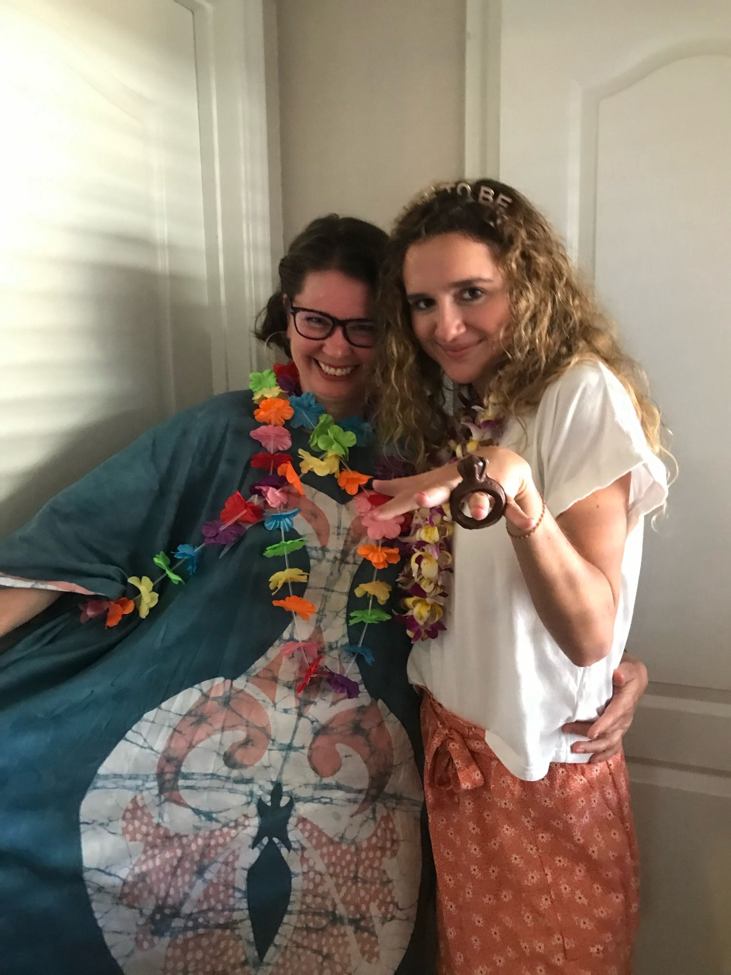 Two pale skinned women, standing in a half embrace. One is displaying a chocolate donut shaped like a wedding ring on her finger. The other is wearing a blue muumu with a vaguely ovarian/vaginal design on the front.