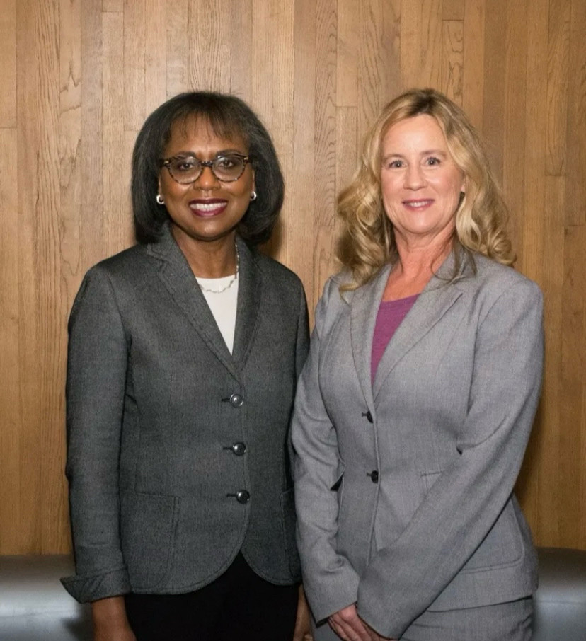 Anita Hill, a Black woman with short dark hair, a bright smile and glasses on her face stands next to Christine Blasey Ford, a white woman with long, wavy blonde hair and a warm smile. Both women are wearing gray jackets with front buttons. Seattle Times