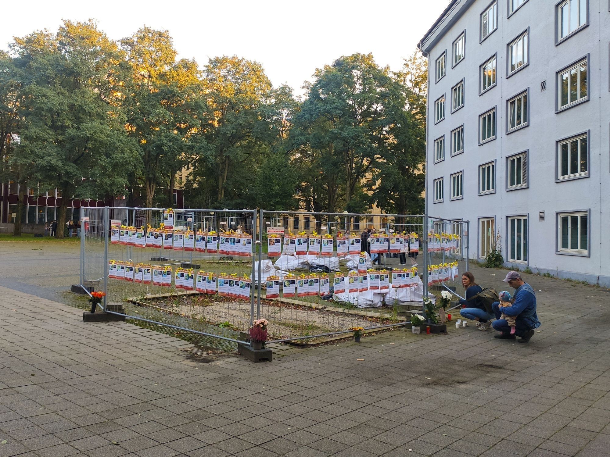 Foto zeigt die Fotos der von Hamas entführeten und ermordeten Personen an einem Bauzaun. Davir Blumen und Kerzen.