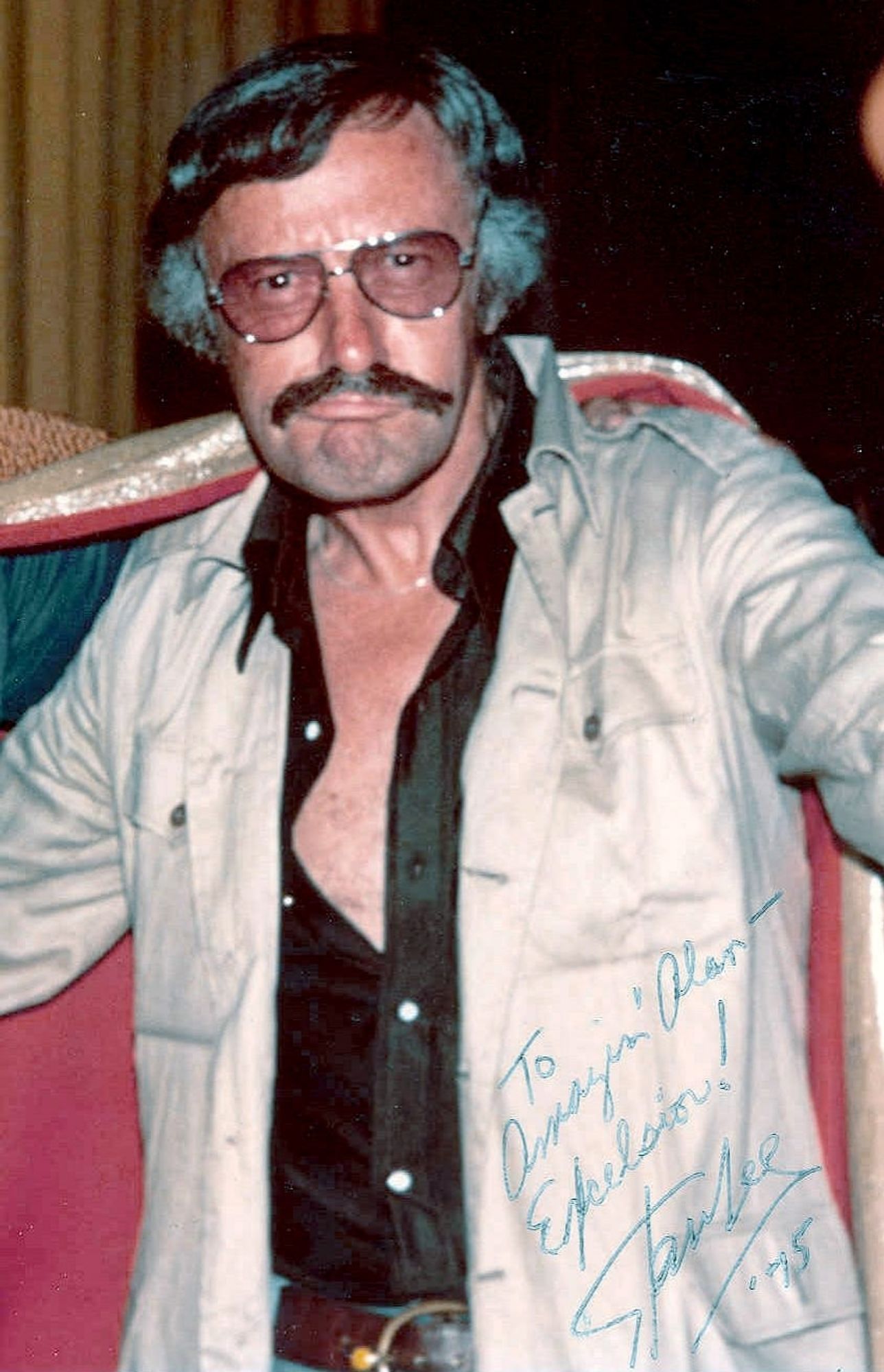 Signed photo of 1970s Stan Lee in a leisure suit jacket over a half-unbuttoned black shirt, with his trademark mustache and aviator sunglasses.