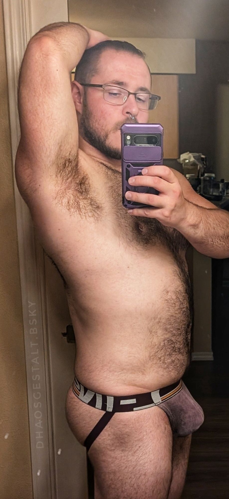 A hairy individual in a burgundy Cin2 jockstrap and glasses, holding a purple phone