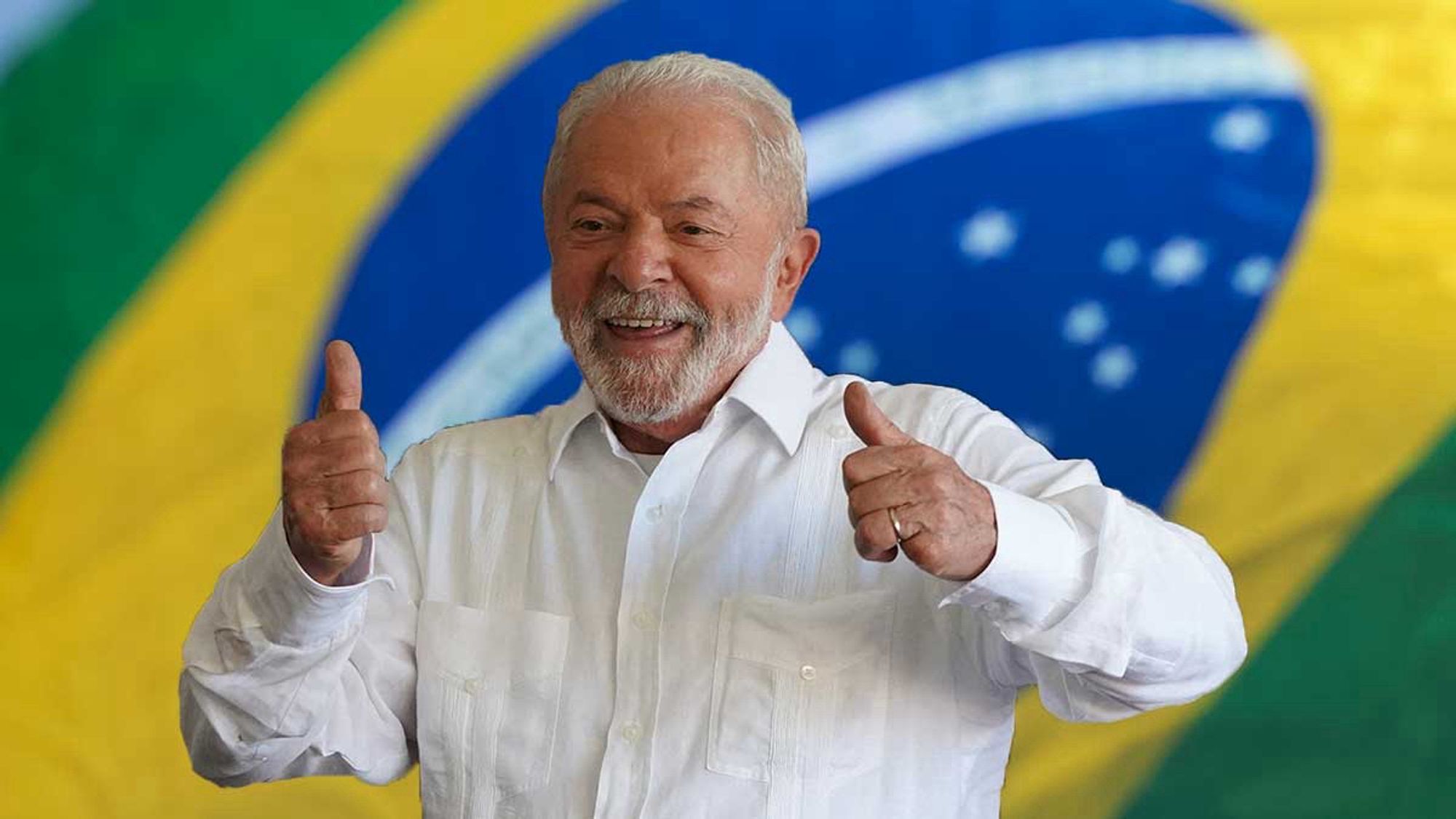 Brazilian President Luiz Inácio Lula da Silva giving thumbs up standing in front of a Brazilian flag