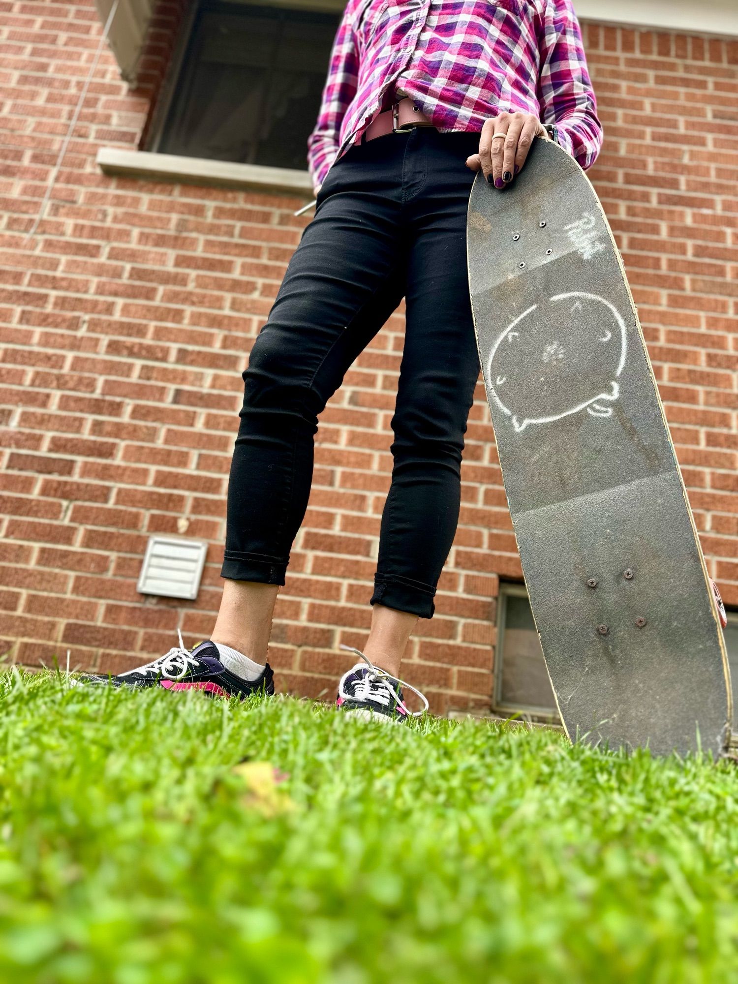 A very low angle shot from the grass of daffodils lower half clad in tight black denim flannel shirt, cute vans skate shoes with a pink stripe. She is holding a long skateboard. this time, grip tape side exposed with a white stencil of a very large round cat that she and her now ex-wife😿 concocted at one point she might bring back and steal as her own don’t tell my wife.