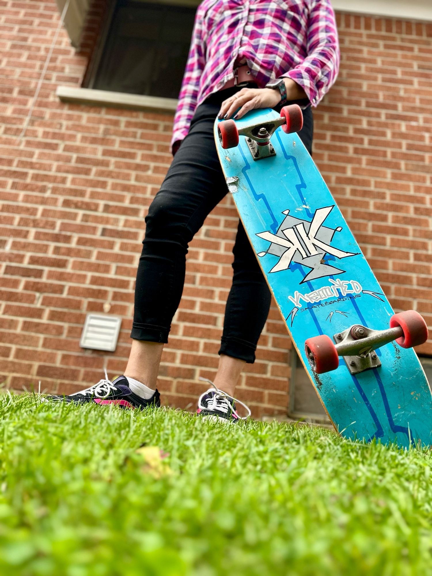 A very low angle shot from the grass of daffodils lower half clad in tight black denim flannel shirt, cute vans skate shoes with a pink stripe. She is holding a long skateboard. Under side of which is teal colored and has a back to back doube K logo and the words Krooked