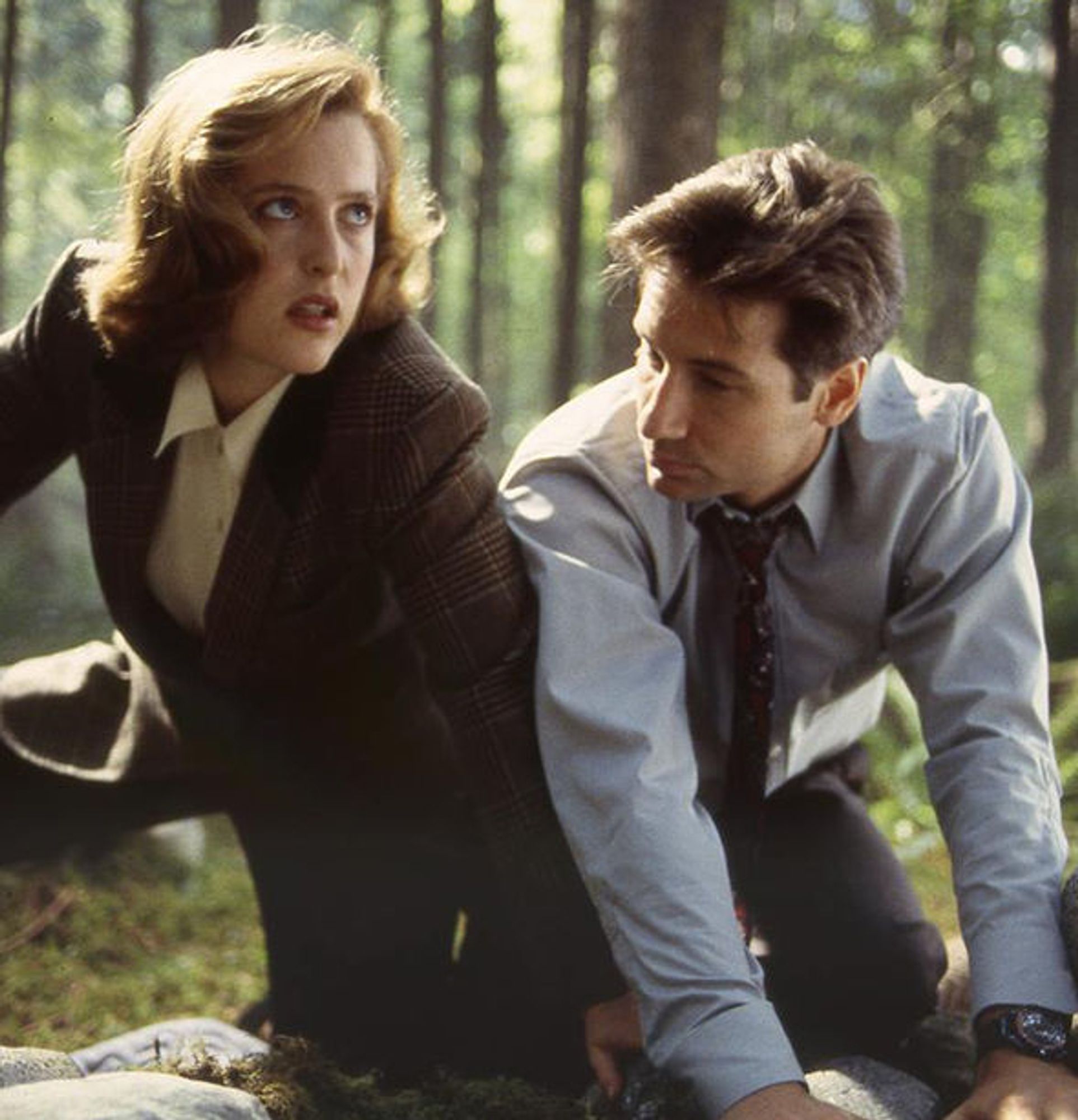 A screencap of Scully and Mulder from The X-Files, probably around Season 2. They're kneeling in a mossy area of a forest near some rocks. She's wearing a brown tweed suit with a cream button-down shirt, looking up and to the left at something offscreen. Mulder has his jacket off, wearing a dark tie and a grey shirt, and his hair is a bit windswept looking. He's looking the opposite direction from Scully, down and to the right. They're close together, their arms touching and Scully's hand resting on the rock between Mulder's hand and his knee.