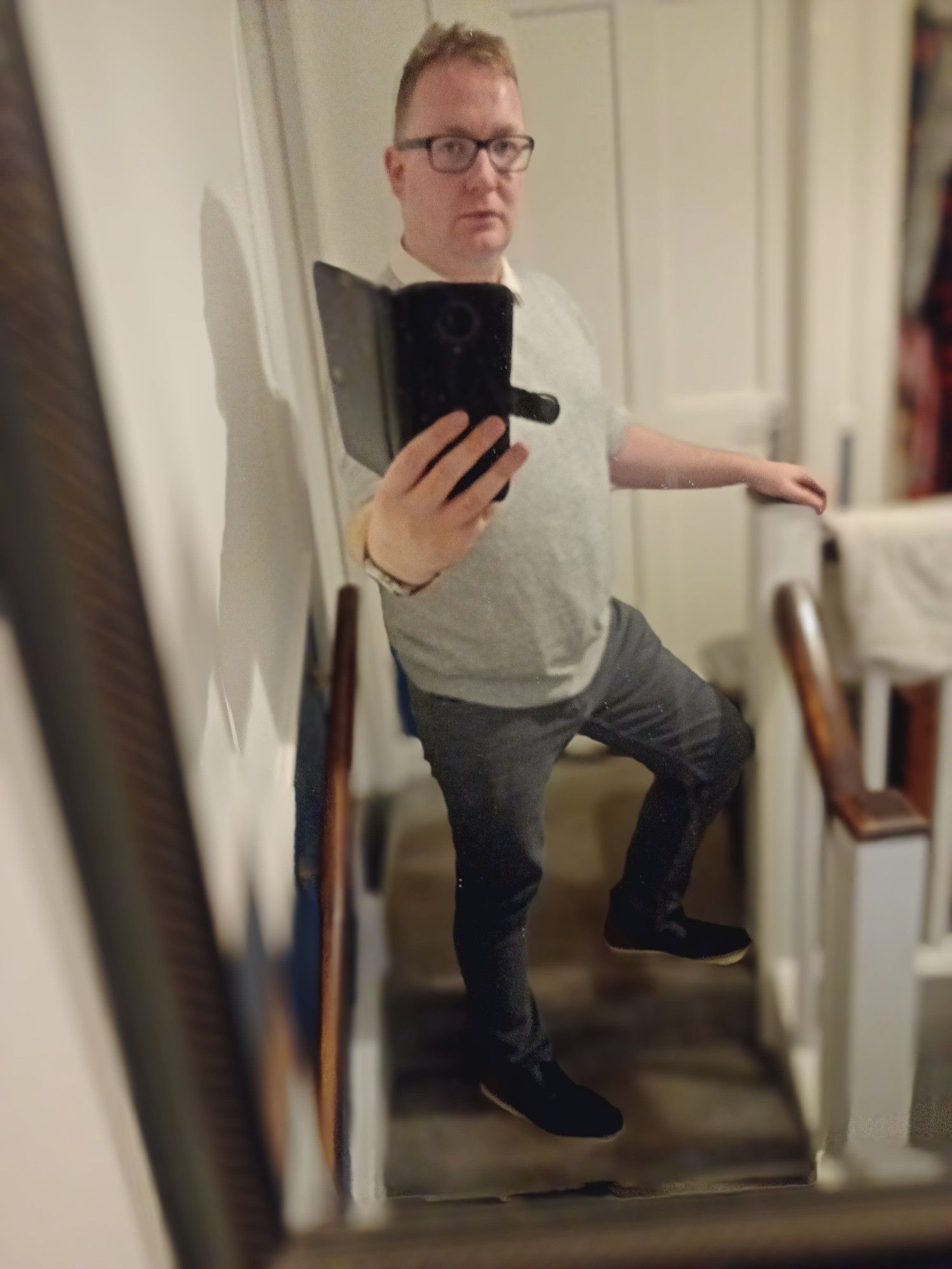 Me, bespectacled white blond non binary person in a grey jumper, dark grey jeans and white collar, standing in a staircase mirror photo