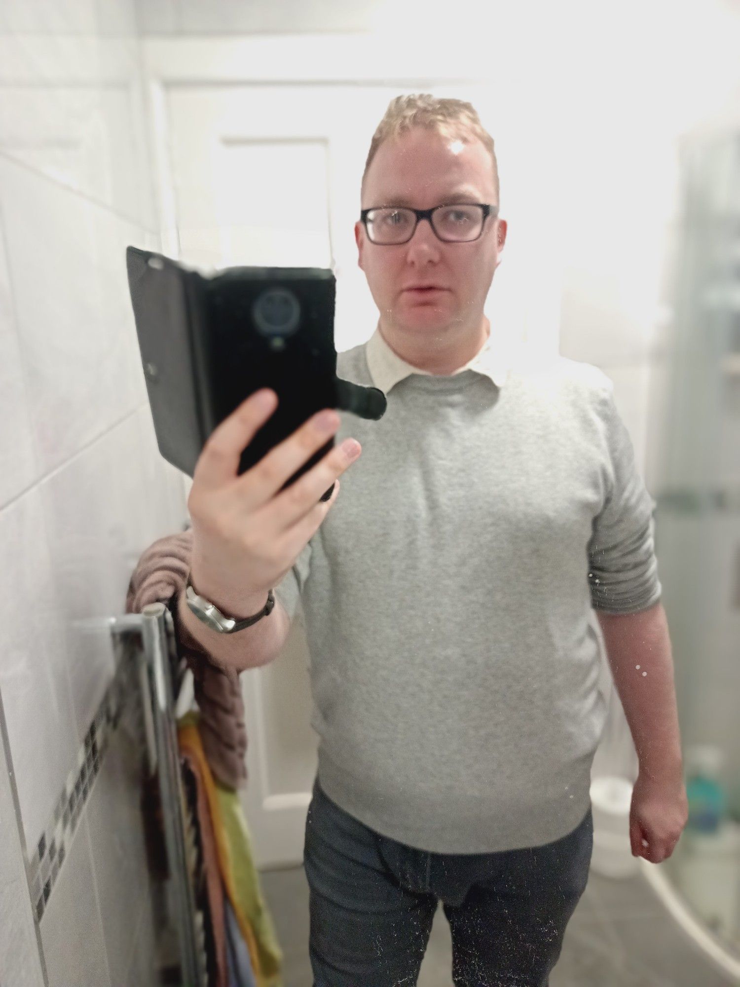 Me, bespectacled white blond non binary person in a grey jumper, dark grey jeans and white collar, standing in a bathroom mirror photo