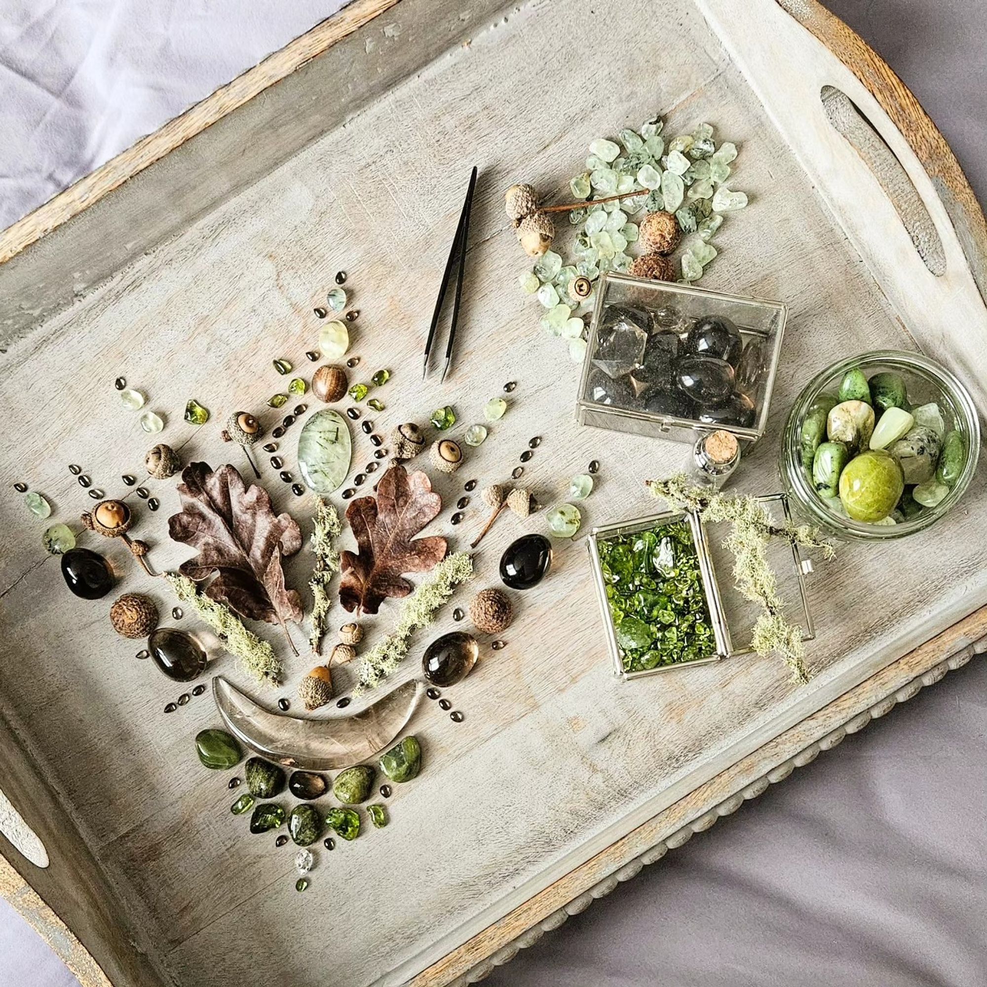 The pale wood tray I use to make my crystal grids on with the above grid created on it, with two glass boxes containing smoky quartz, and peridot, and a glass circular bowl full of green crystal stones, and a few twigs covered in reindeer moss. And my tweezers, which I use to place the tiny gems!