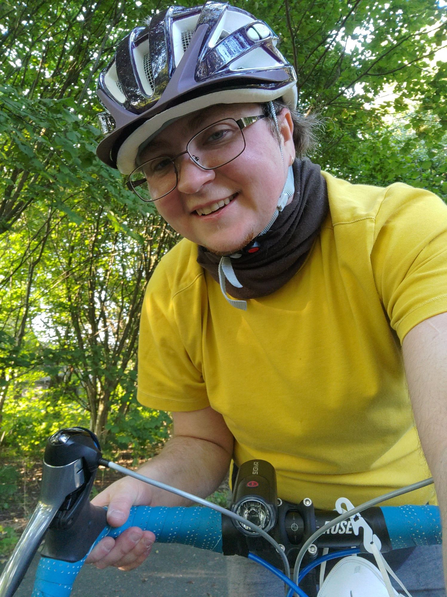 Ein skye auf einem Rennrad macht ein Selfie über den Lenker. Ich bin klein, weiß, hab kurze Haare, wirke eher androgyn und trage einen Fahrradhelm und ein breites zufriedenes Lächeln.