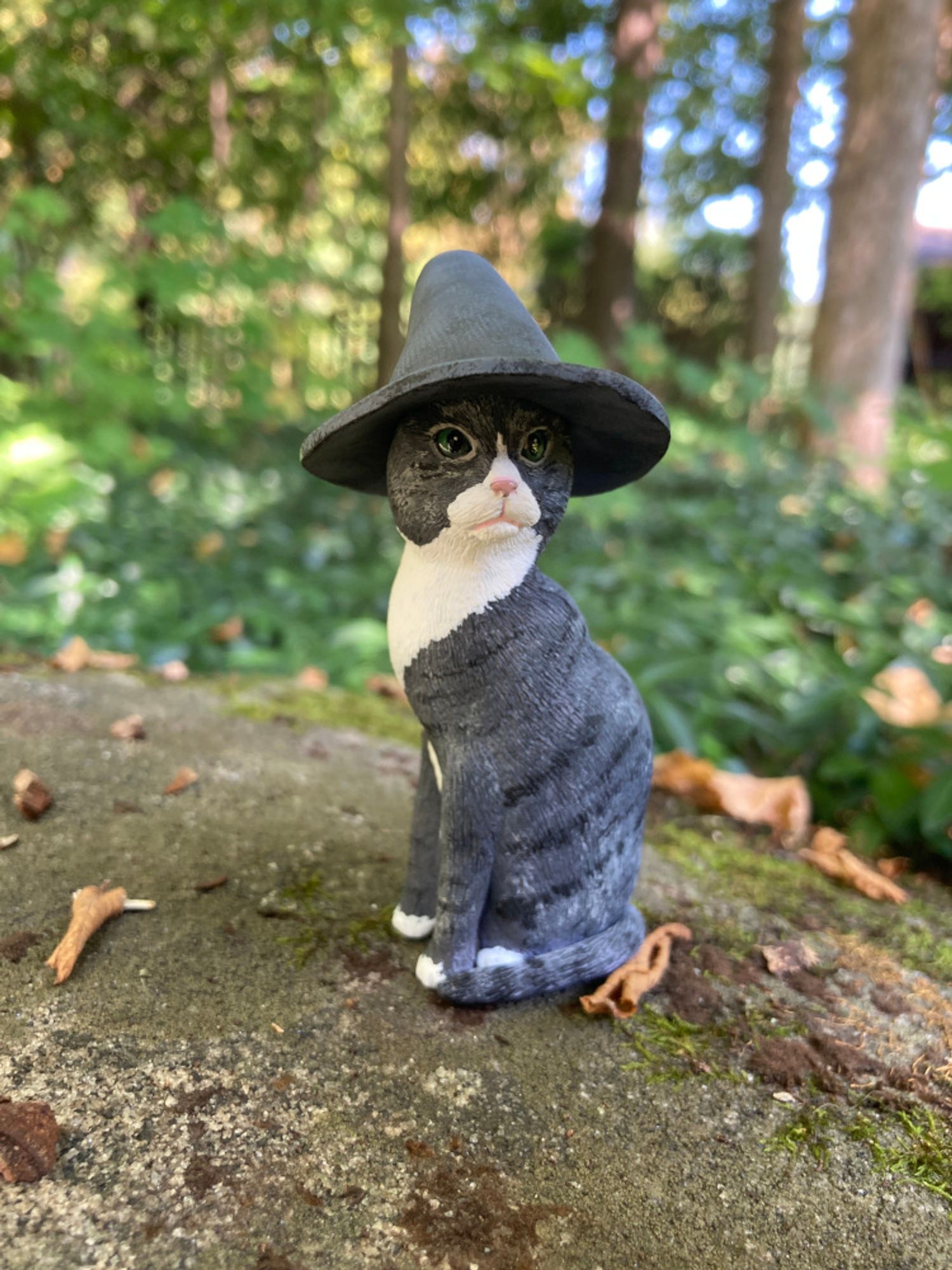 Clay sculpture of a grey tabby cat with green eyes seated on a rock in a forest. He is wearing a large grey wizard's hat and staring off into the distance.