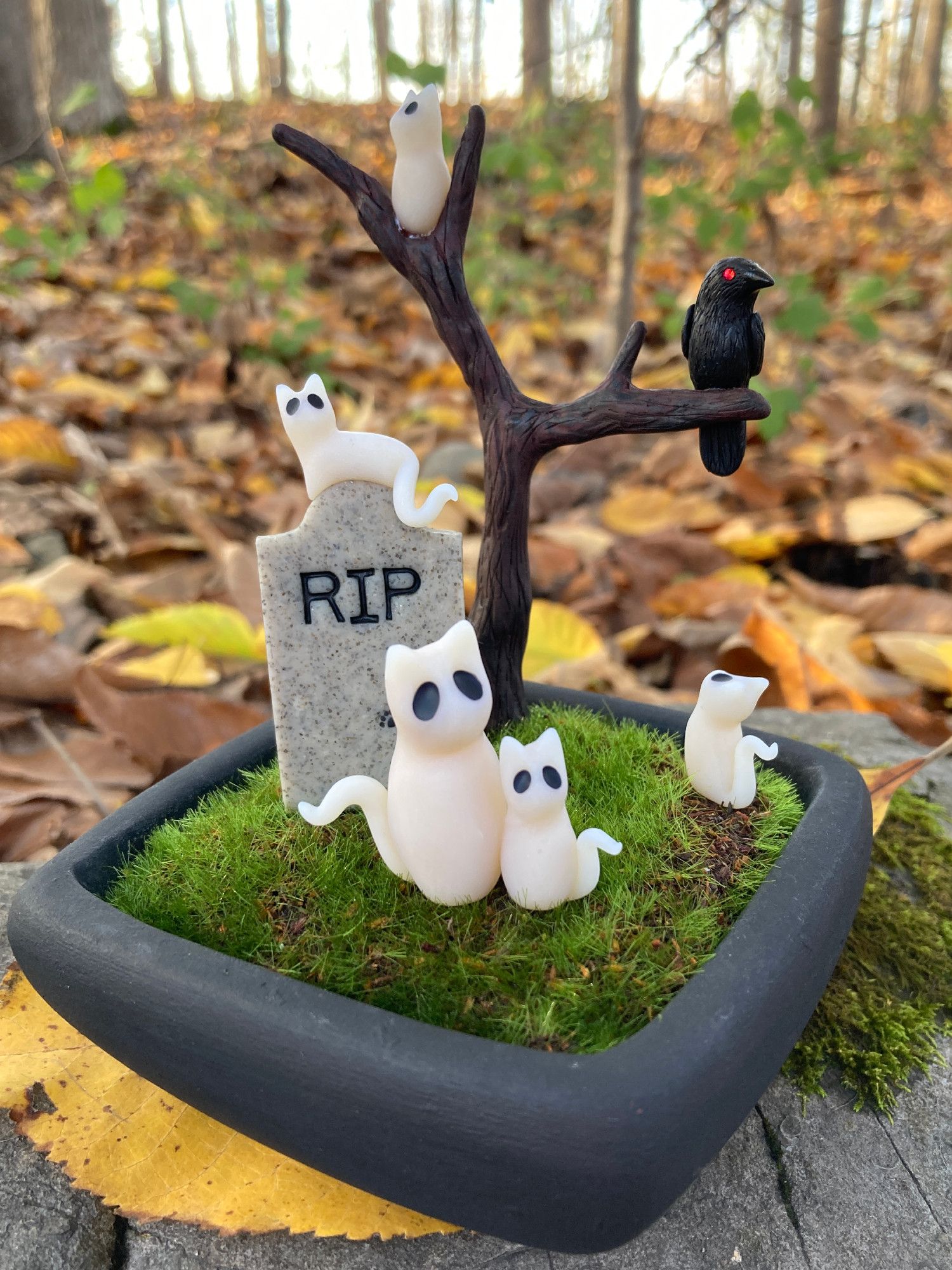 Clay sculpture of several ghost cats - a mother and four kittens - in a small graveyard with a head stone and leafless tree with a raven. The sculpture sits in a black ceramic pot.