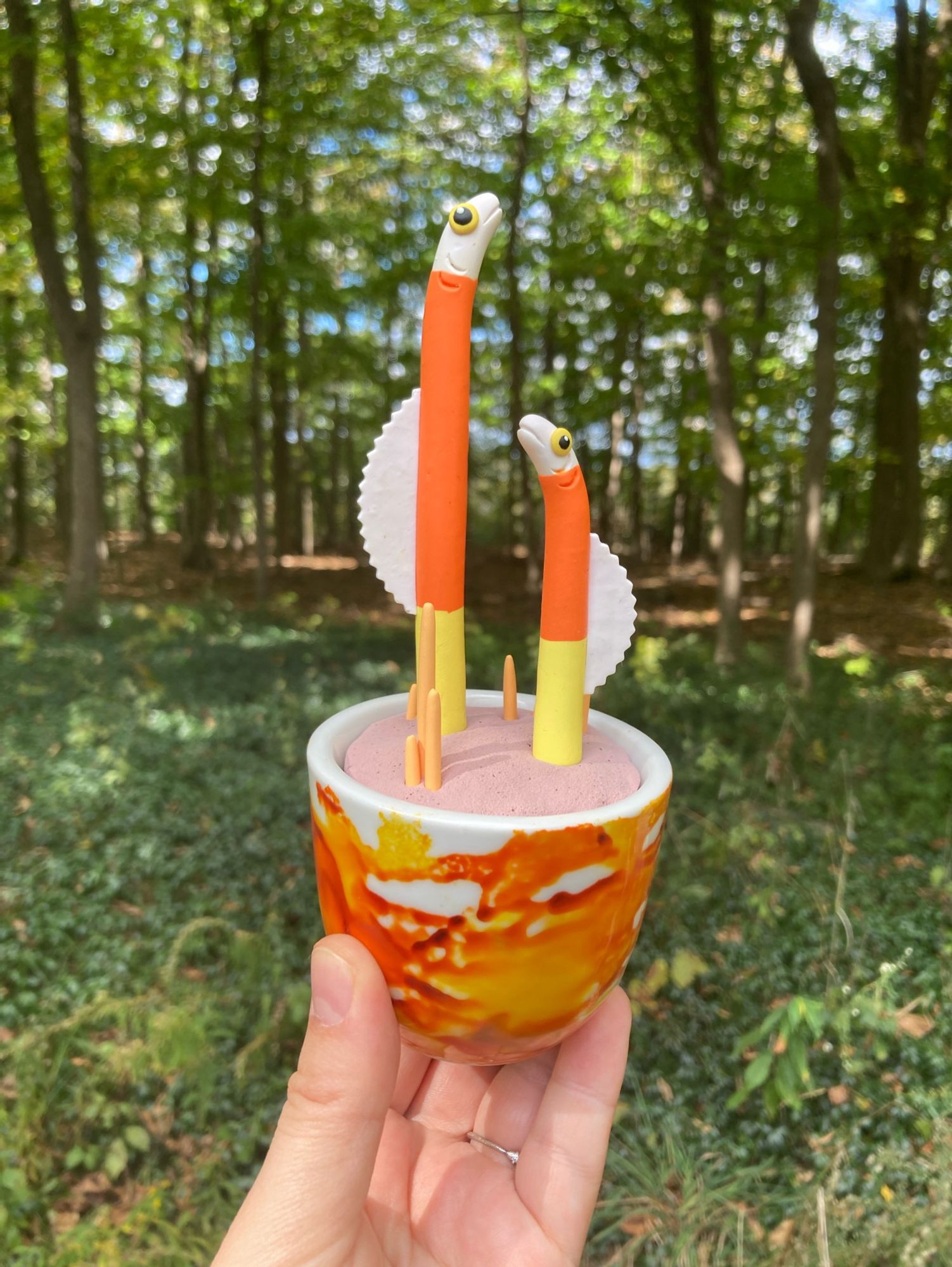 Clay sculpture of two eels, colored like candy corn, “planted” in clay sand with bright orange coral in a tie-dyed orange, yellow, and white pot.