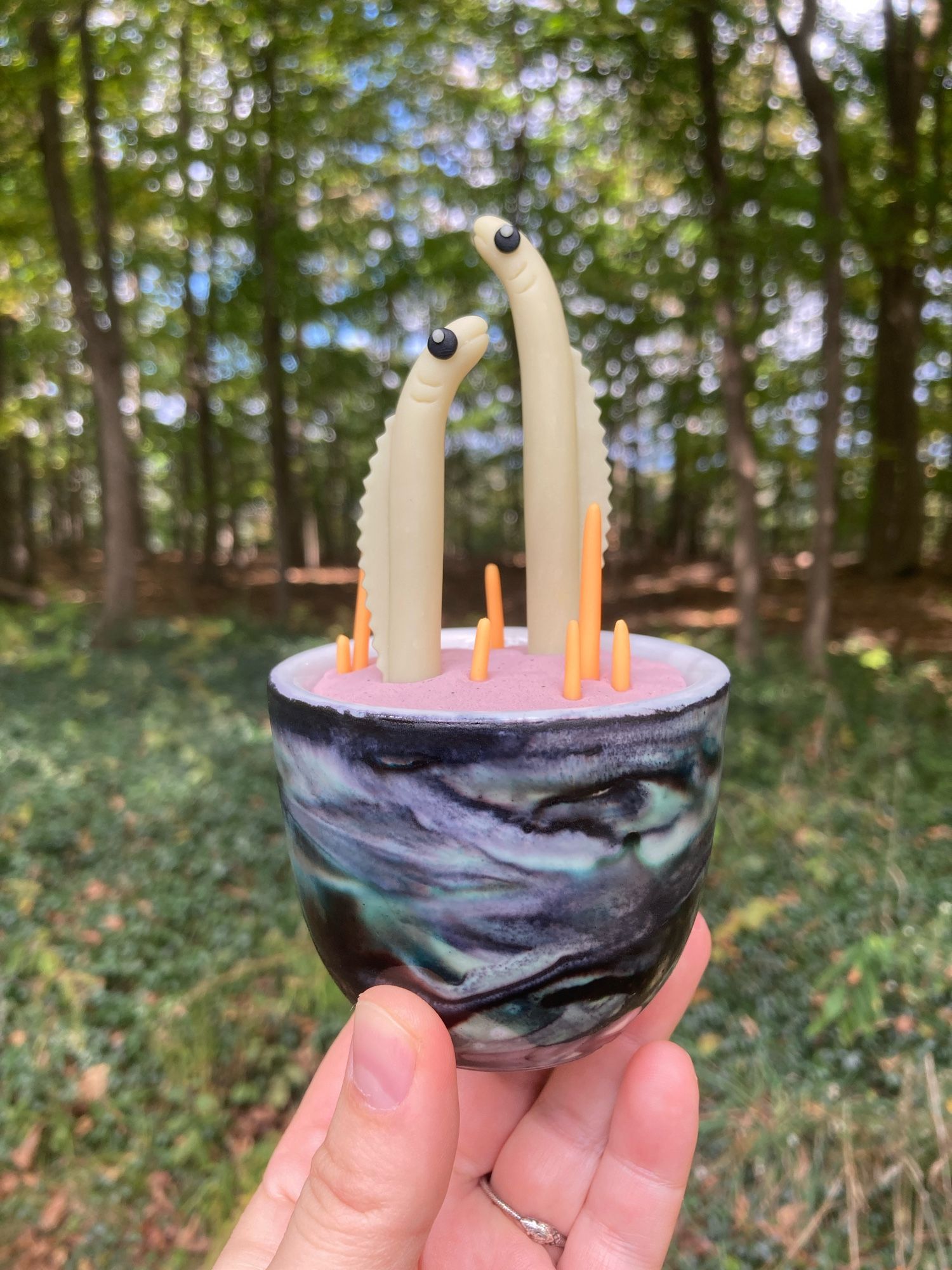 Clay sculpture of two pale white eels with black eyes, “planted” in clay sand with bright orange coral in a black, purple, and green tie-dyed pot.