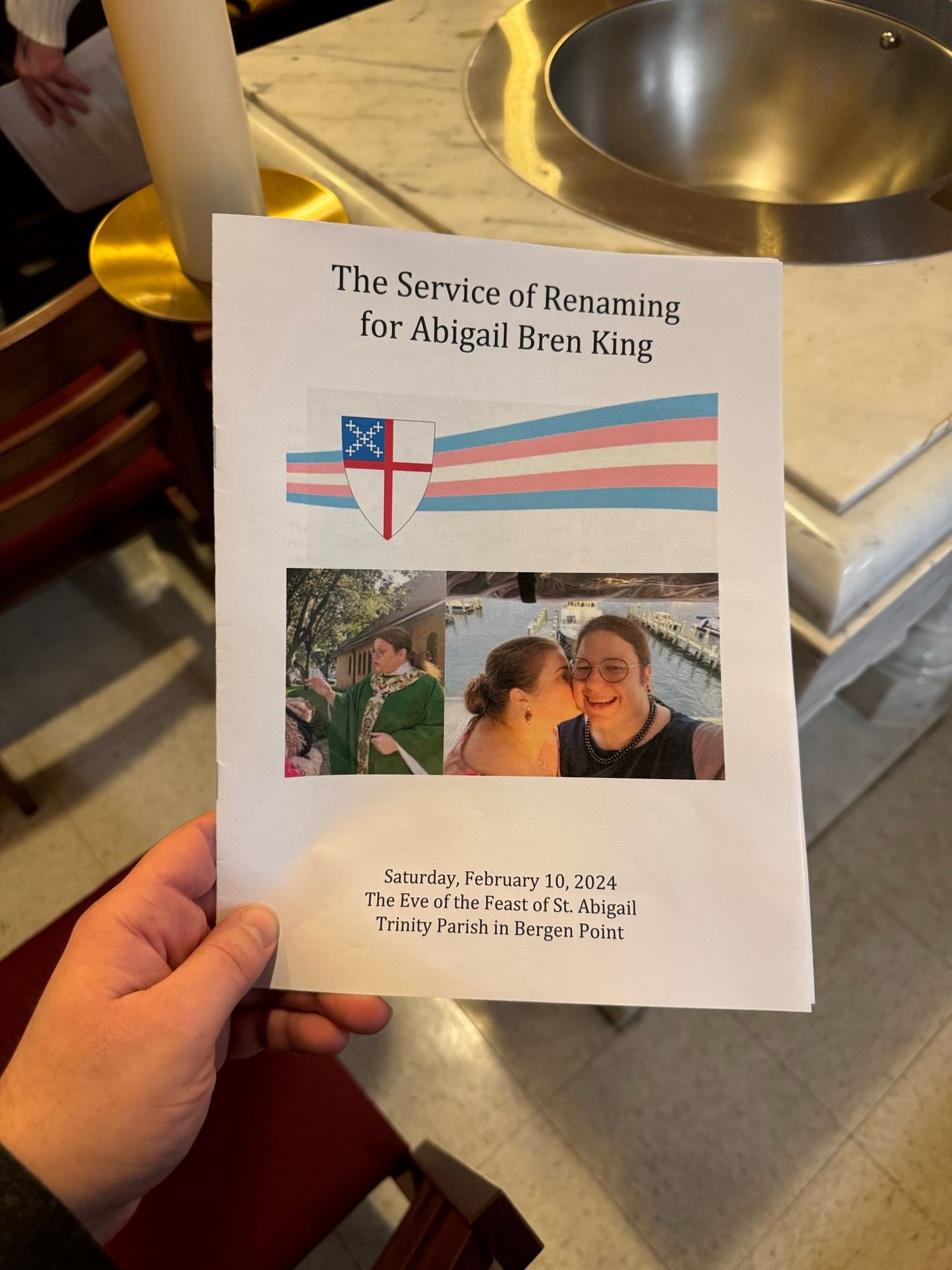 Service leaflet in front of a baptismal font and beside a Paschal Candle. Cover says “The Service of Renaming for Abigail Bren King” and “Saturday, February 10, 2024 The Eve of the Feast of St. Abigail Trinity Parish in Bergen Point”. The cover has a picture of Abigail in priest vestments and a picture of Abigail and her wife. There is also a graphic with the Trans Pride Flag with the Episcopal Shield in front of it.