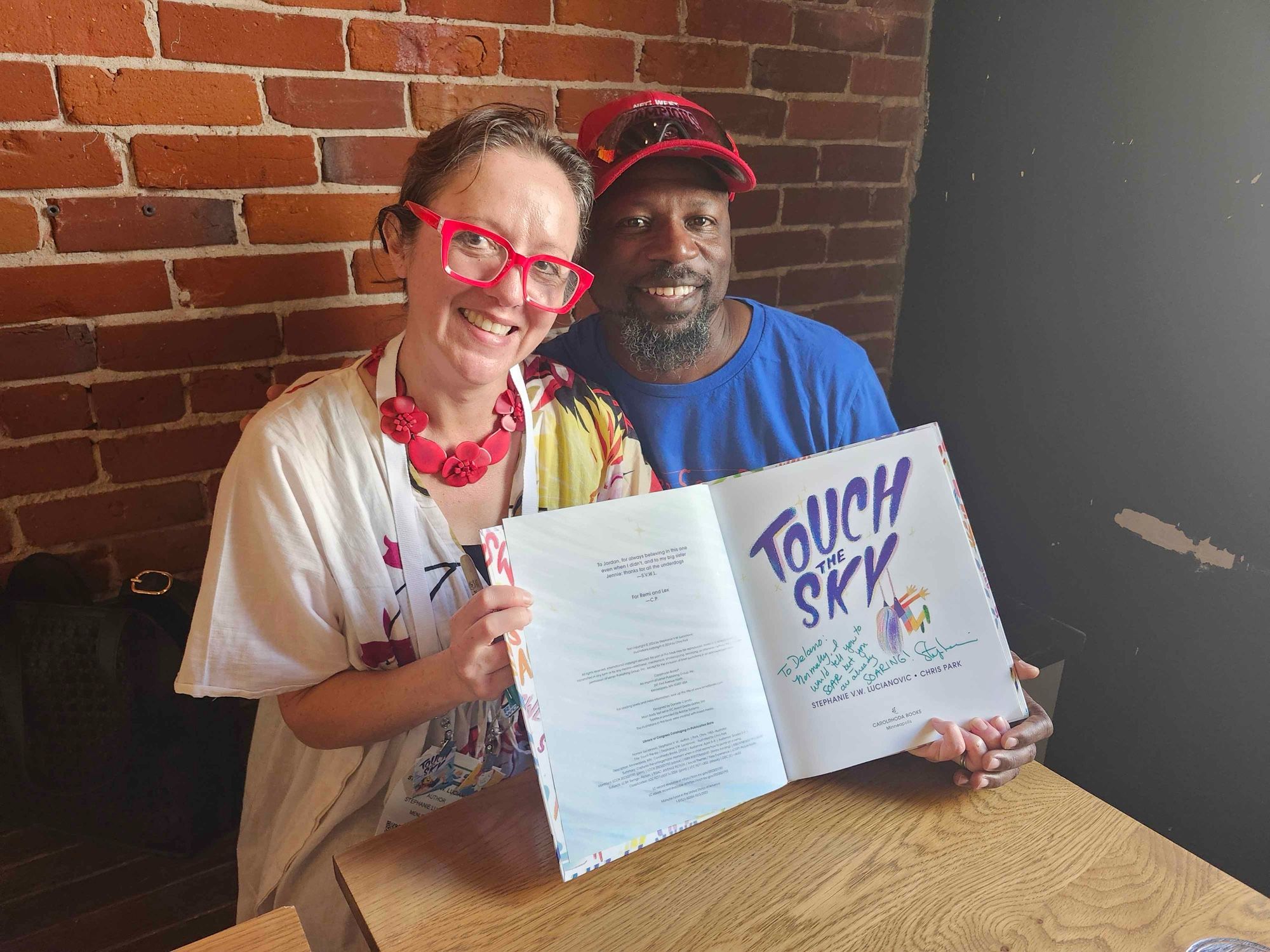 Stephanie in red glasses and Delano in a red hat and blue shirt holding an open signed copy of TOUCH THE SKY