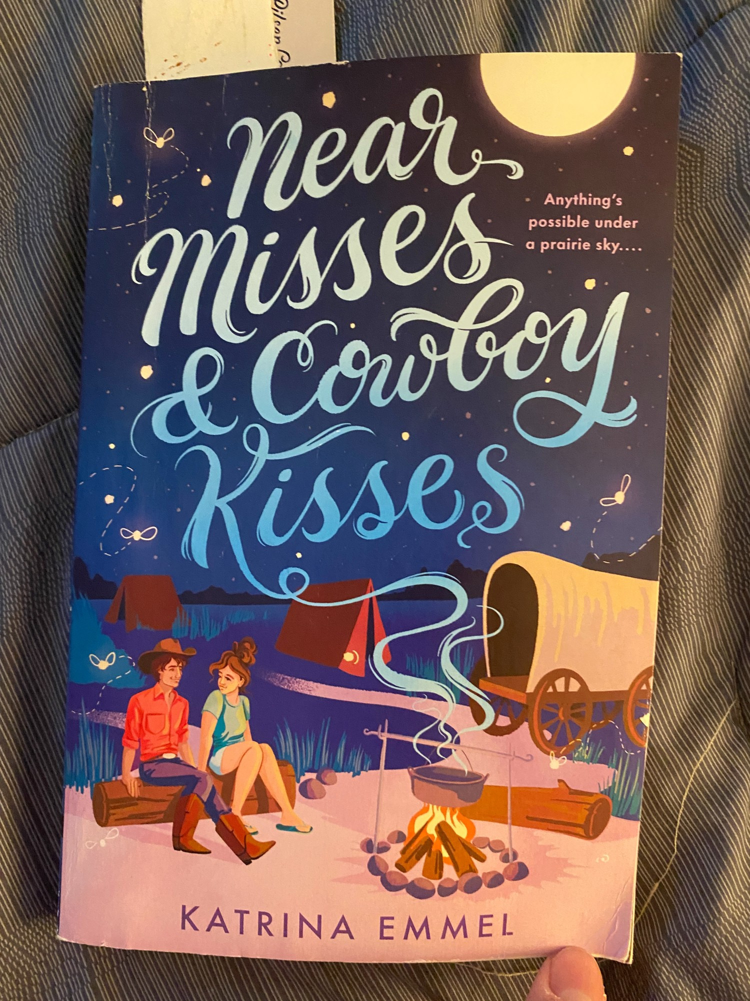 Cover of the YA novel Near Misses and Cowboy Kisses by Katrina Emmel. Cover art features two teens (boy and girl-presenting) sitting on a log in front of a campfire, under a moon and dark sky, with tents and a covered wagon nearby.