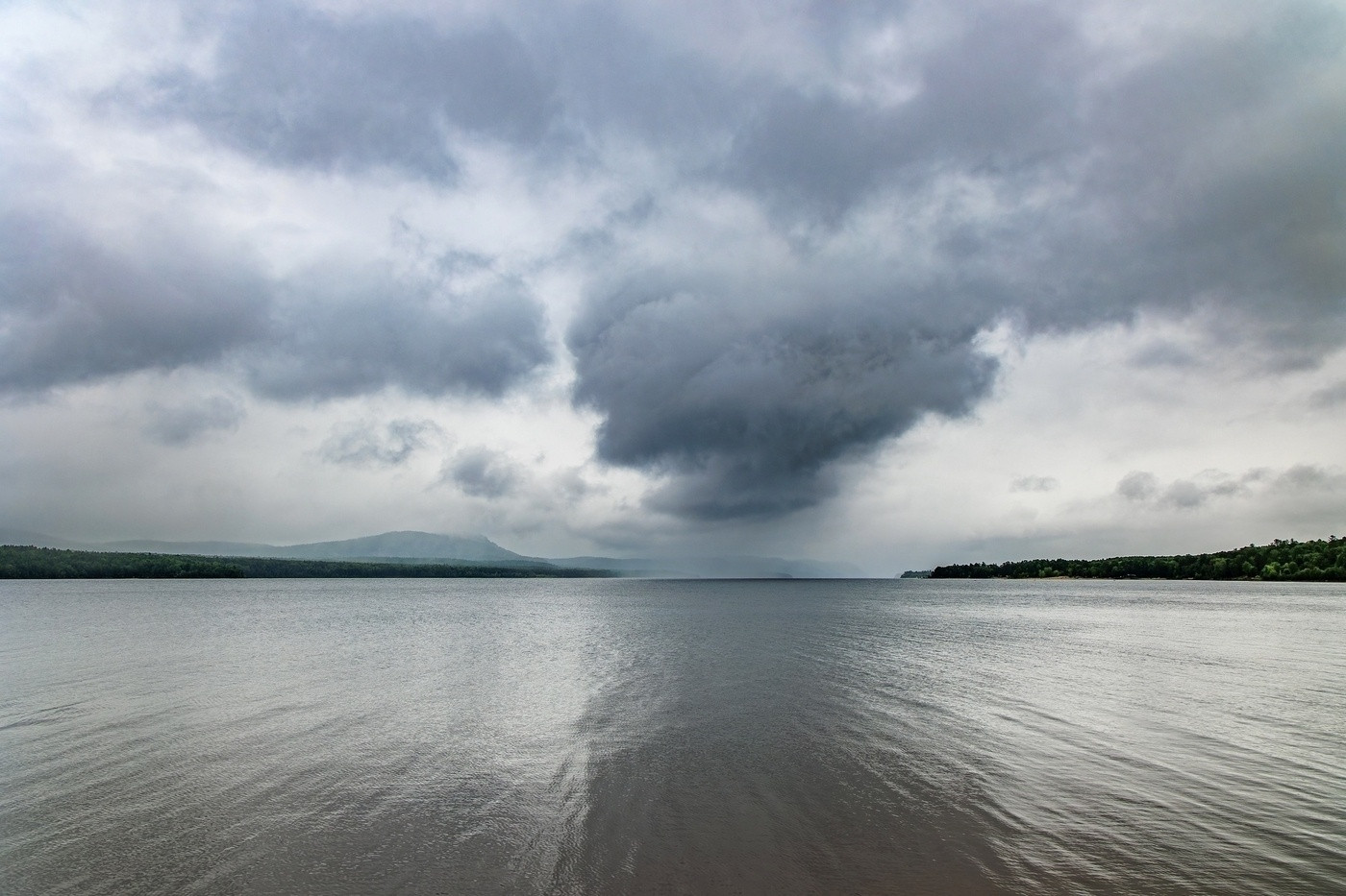 A wide, tranquil river stretches into the distance under a dramatic, cloud-filled sky. Mount Martin is on the far side of the water.