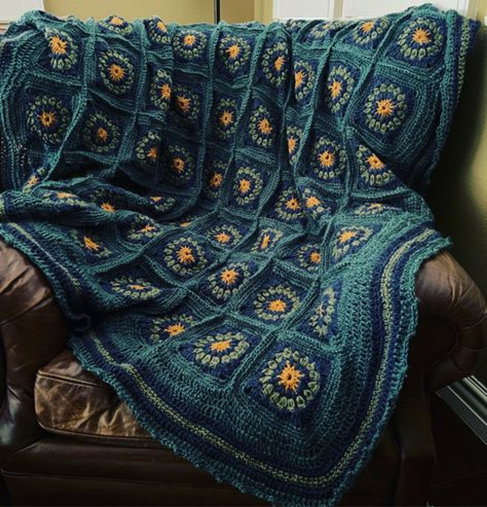 A crochet granny square blanket in golden yellow and shades of blue, draped over a battered leather armchair.