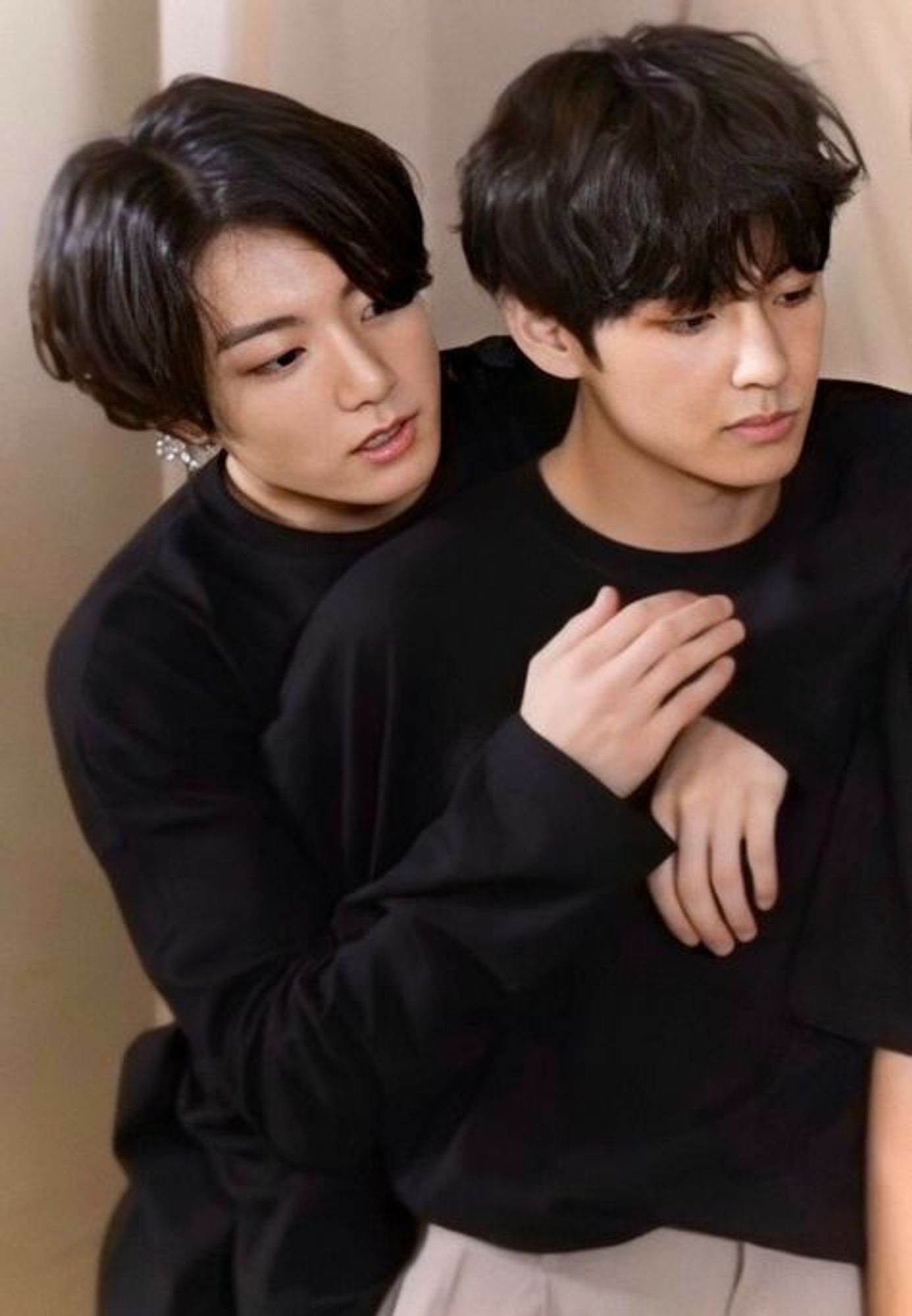 Jungkook and Taehyung, both with floppy black hair and black sweatshirts, looking at something off-camera. Jungkook is draped over Tae's back and looks a little apprehensive.