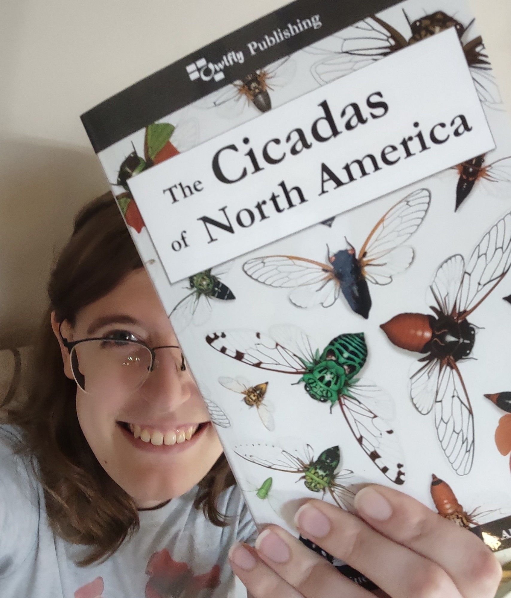 A twenty-something woman with brown hair and glasses holds up a book she wrote titled "The Cicadas of North America" in the foreground. The cover design shows illustrations of cicadas swirling outwards from a central point.