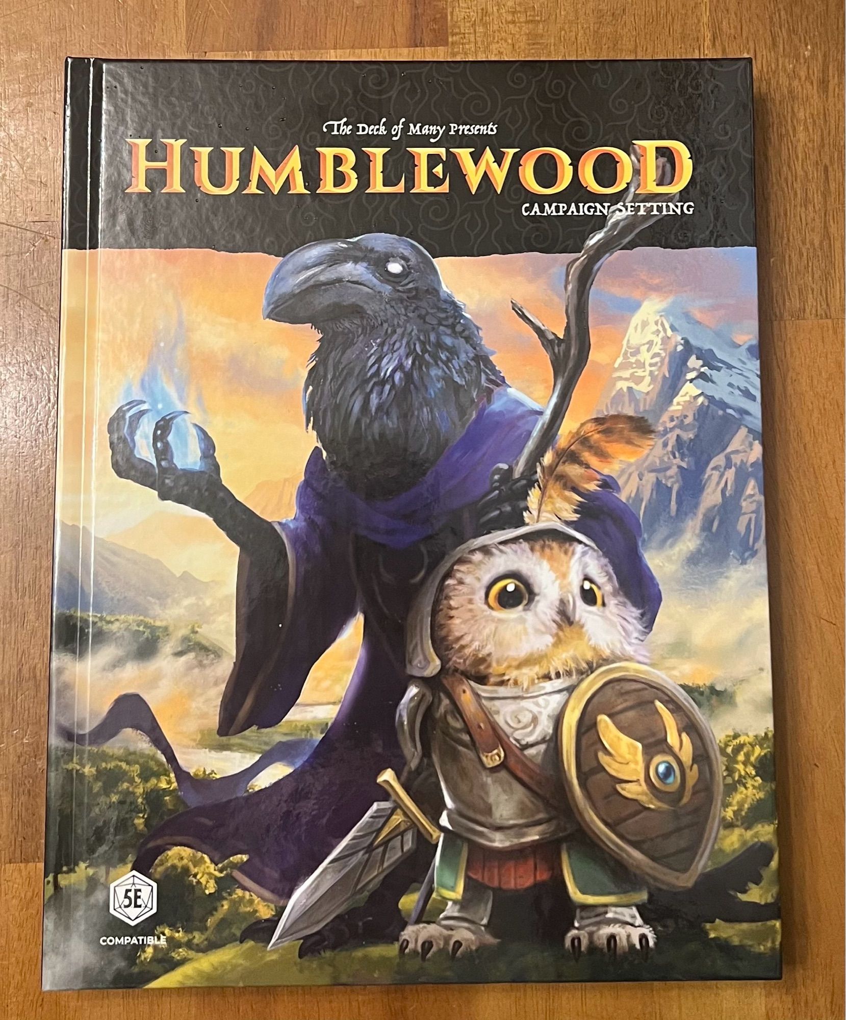 The hardcover of the Humblewood Campaign Setting

Pictured are a crow like humanoid in purple robes, casting a spell with his right hand and holding a wizards staff in his left and in front of him a little brown and white owl person dressed in armor and  holding a sword and shield.