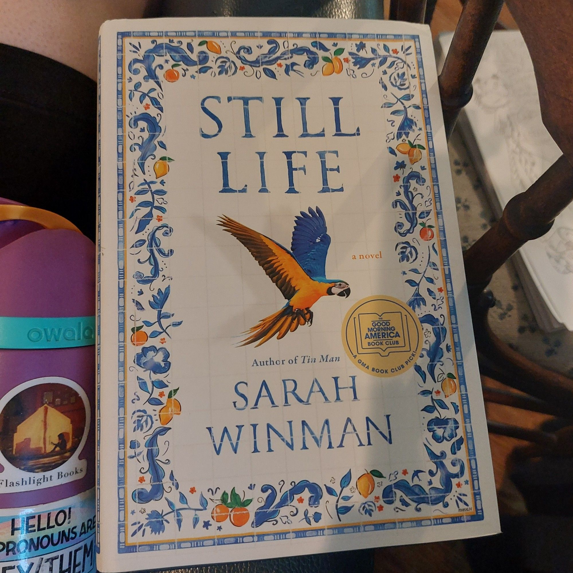 Photo of the hardcover edition of Still Life by Sarah Winman. Cover depicts a blue and yellow parrot on a white background. Border is done to look like tiles with blue designs and yellow fruit.