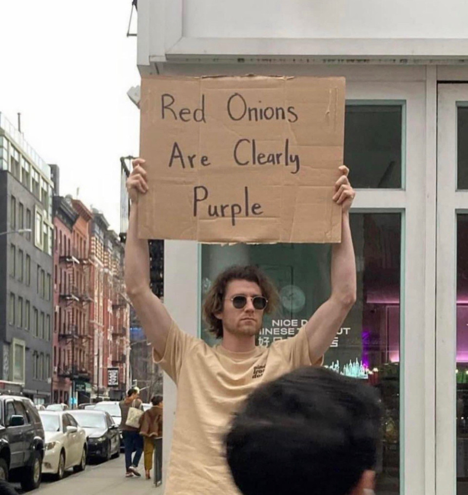 A man on a street holding a sign that says red onions are clearly purple