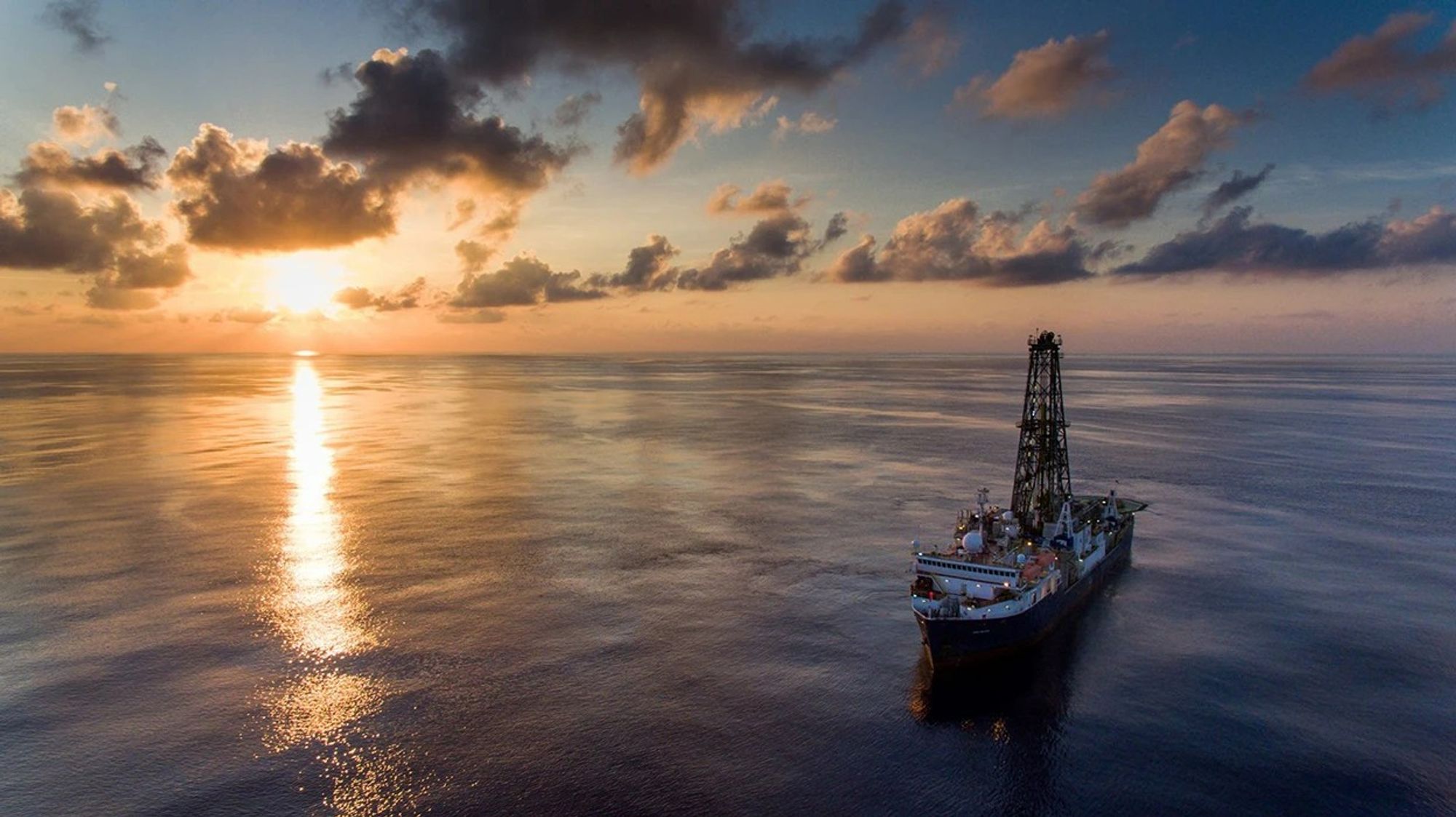 Sunset over the ocean with a a ship in the foreground that has a drill derrick on it (the "JOIDES Resolution")