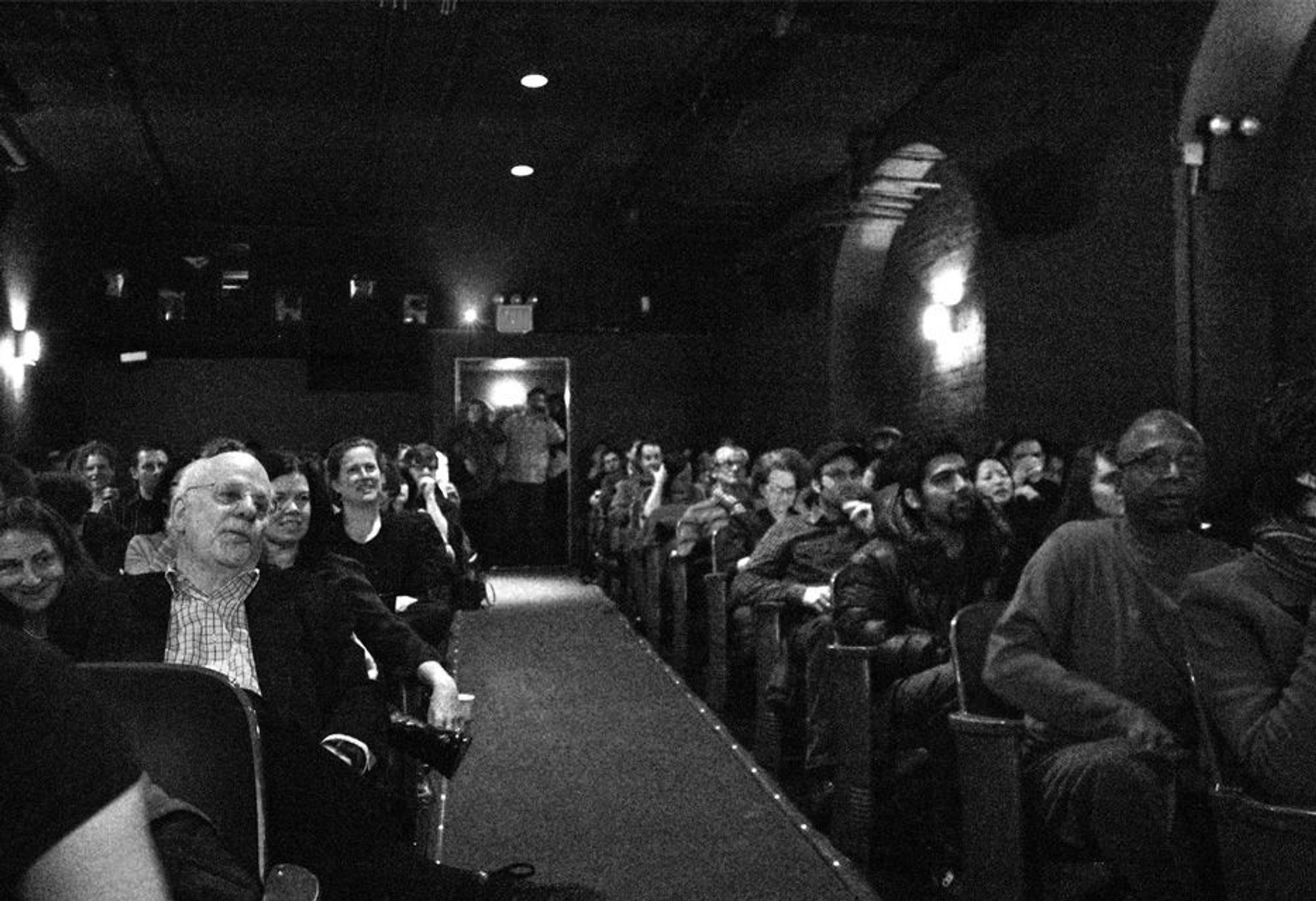 A cinema audience. The photo is taken from where the screen would be. The photo is black and white. Dozens of faces are visible, all showing different emotions