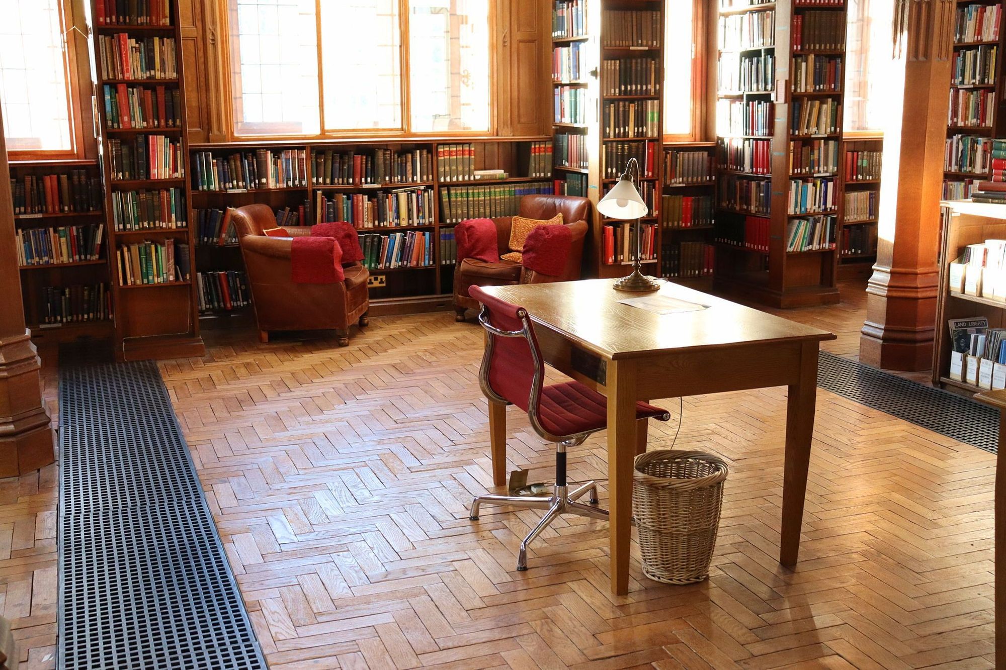 A desk in the Theology Room. There is a red chair next to it and a lamp atop it. There are bookshelves and windows in the background.