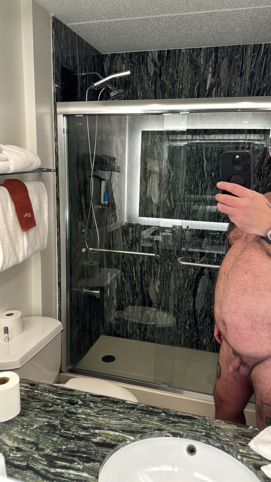 The torso and legs of a naked bear taking a picture of the marbled shower in his hotel room. You can see his soft small cock, but not his face. 