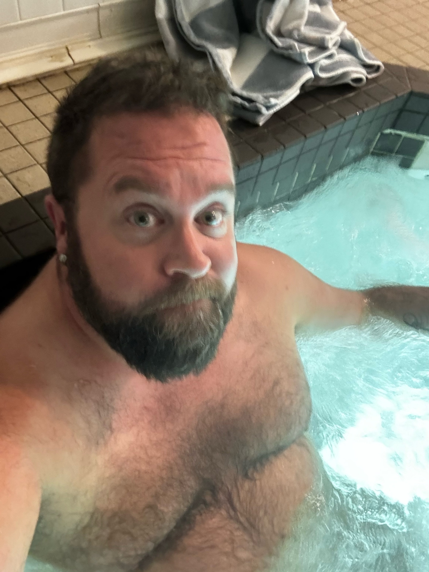 A bear seated in the hotel’s hottub. Not wearing his glasses cuz they’re fogged up; with his pool towel on the tiled floor behind him. 