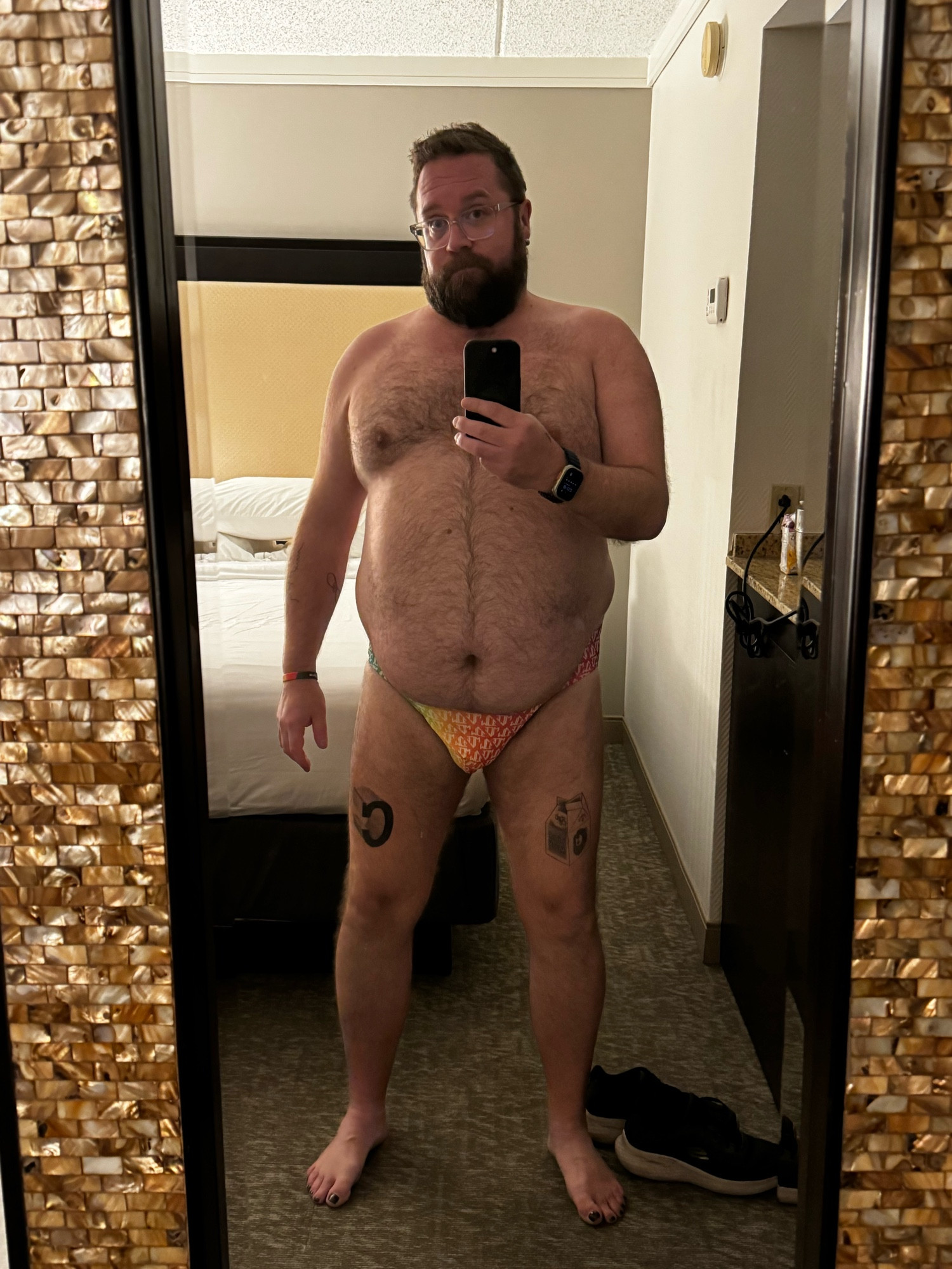 A bear, standing in front of his hotel room full length mirror, taking a selfie wearing his MCE Creations speedo. It is a gradient green, yellow, pink, red color over the pouch of the speedo; in the background is one of his room’s beds. 