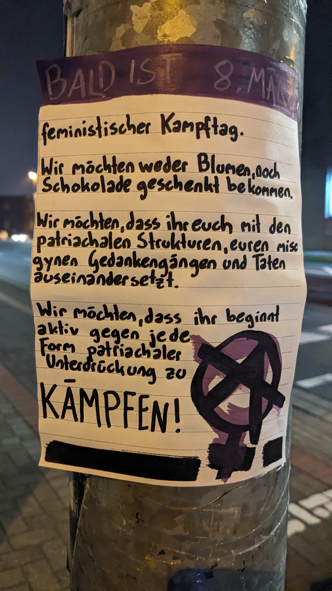 Foto von einem kleinen selbstgeschriebenen Plakat (zwischen A5 und A4 groß) an einem Laternenmast: "Bald ist 8. März, feministischer Kampftag. Wir möchten weder Blumen noch Schokolade geschenkt bekommen. Wir möchten dass ihr euch mit den patriarchalen Strukturen, euren misogynen Gedankengängen und Taten auseinander setzt. Wir möchten dass ihr beginnt aktiv gegen jede Form patriarchaler Unterdrückung zu KÄMPFEN!"
Unten auf dem Plakat ist ein schwarz-lila Anarchafeminismus-Zeichen